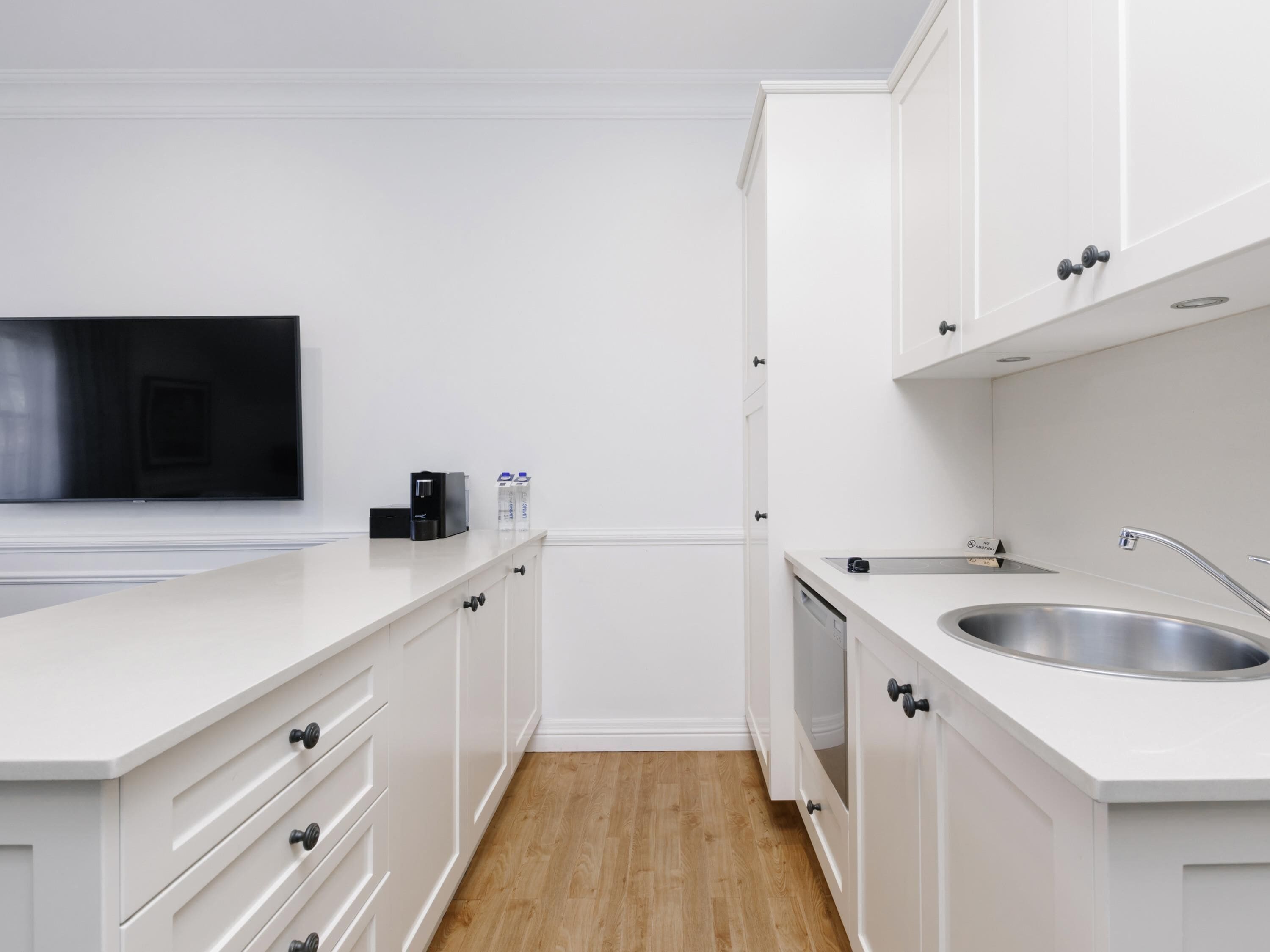 Una cocina moderna con armarios blancos, lavavajillas empotrado, pileta de acero inoxidable y un televisor de pantalla plana montado en la pared.