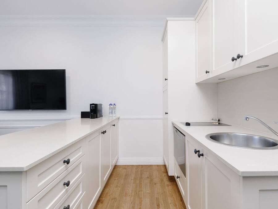 A modern kitchen with white cabinetry, a built-in dishwasher, a stainless steel sink, and a flat-screen TV mounted on the wall.