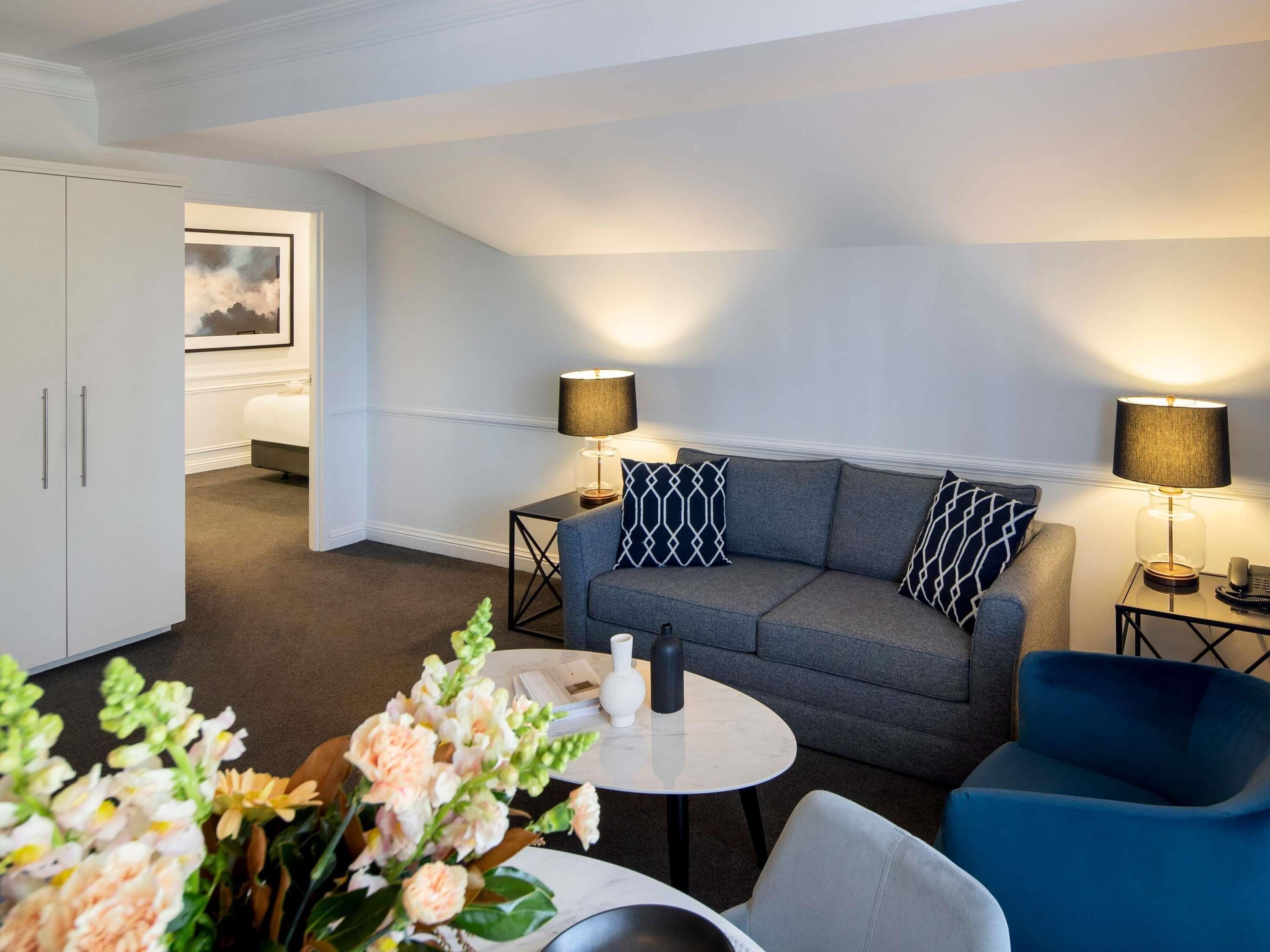 Ein modernes Wohnzimmer mit einem grauen Sofa, zwei Beistelltischen mit Lampen, einem weißen Schrank und einem kleinen runden Tisch mit einer Blumenvase.
