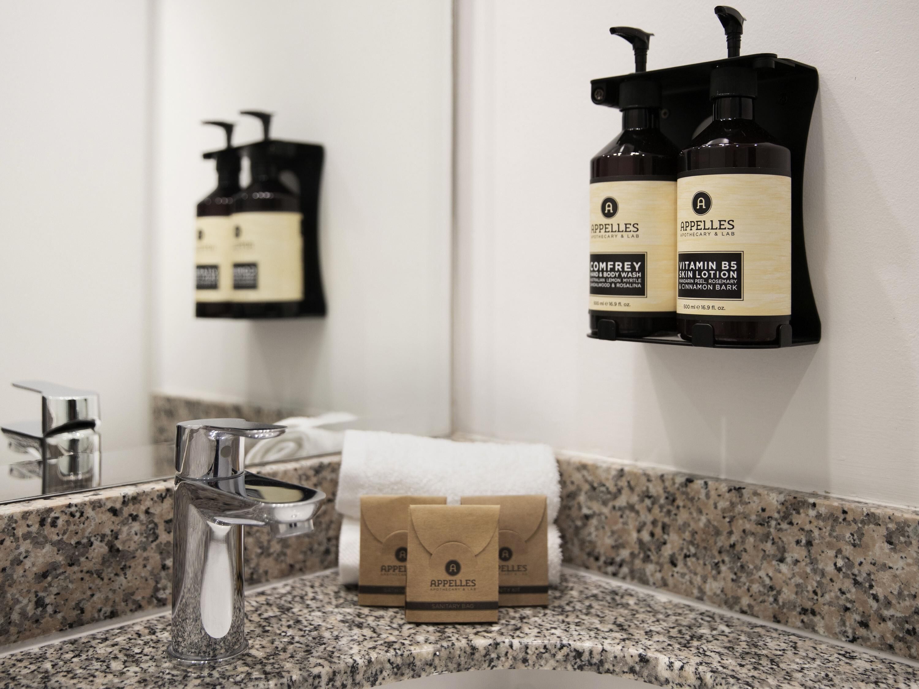 A bathroom with a granite countertop, a mirror, a sink, and two bottles of Apelles body wash on a wall-mounted dispenser.