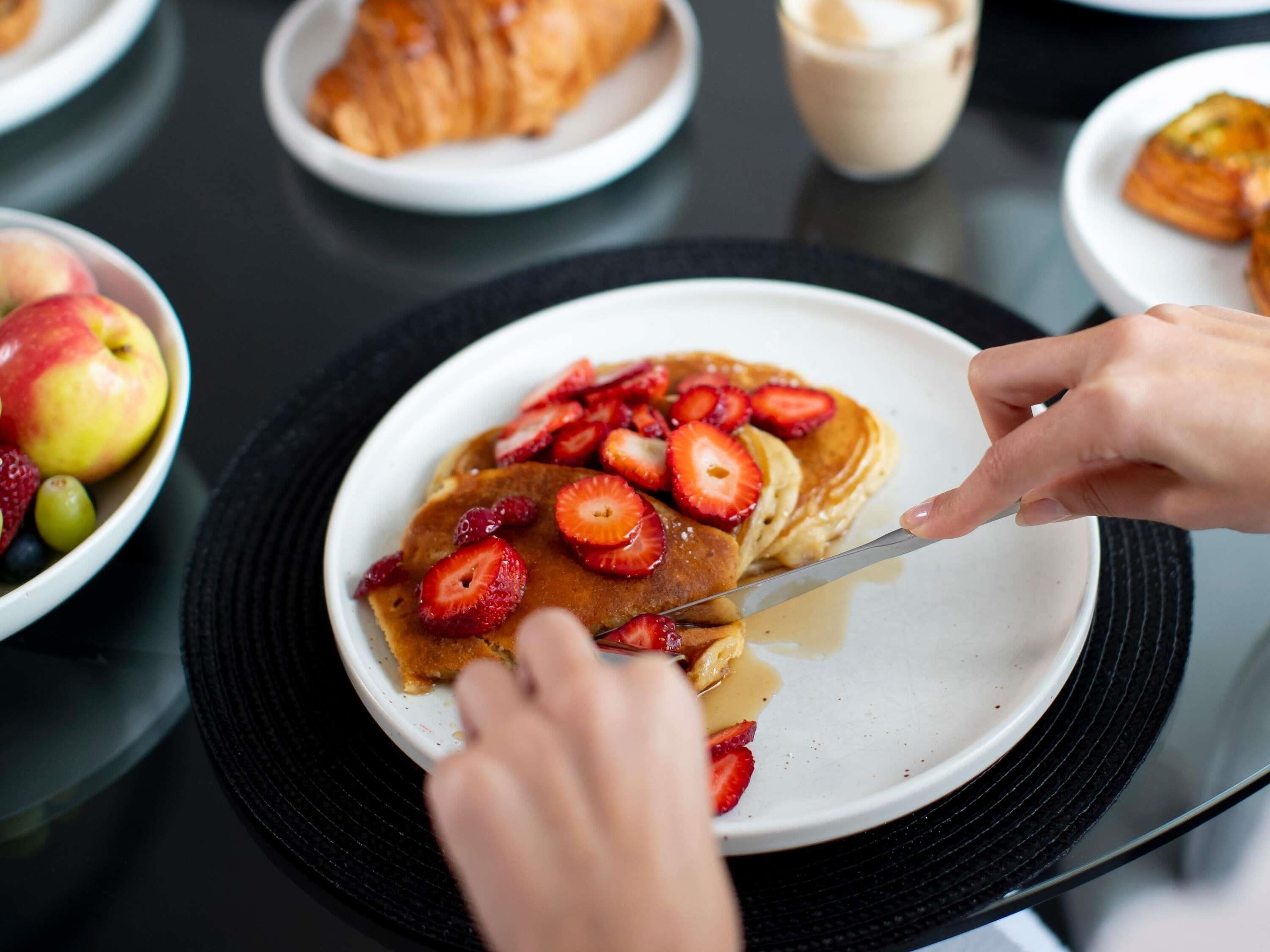 A breakfast setting with pancakes topped with strawberries and syrup, a croissant, and a bowl of fruit.