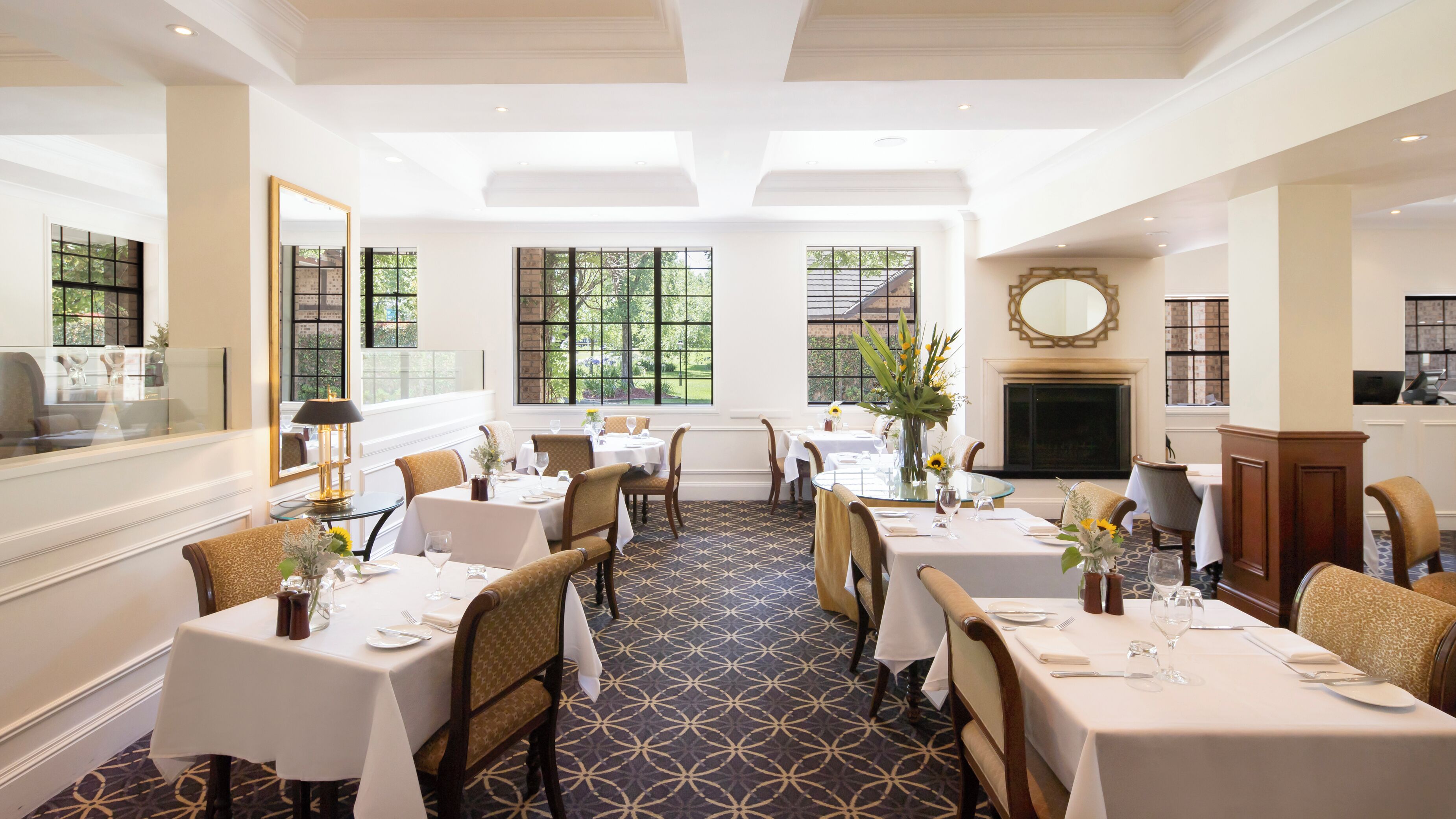 A dining area with multiple tables set for guests, large windows offering views of the outdoors, and a fireplace in the background.