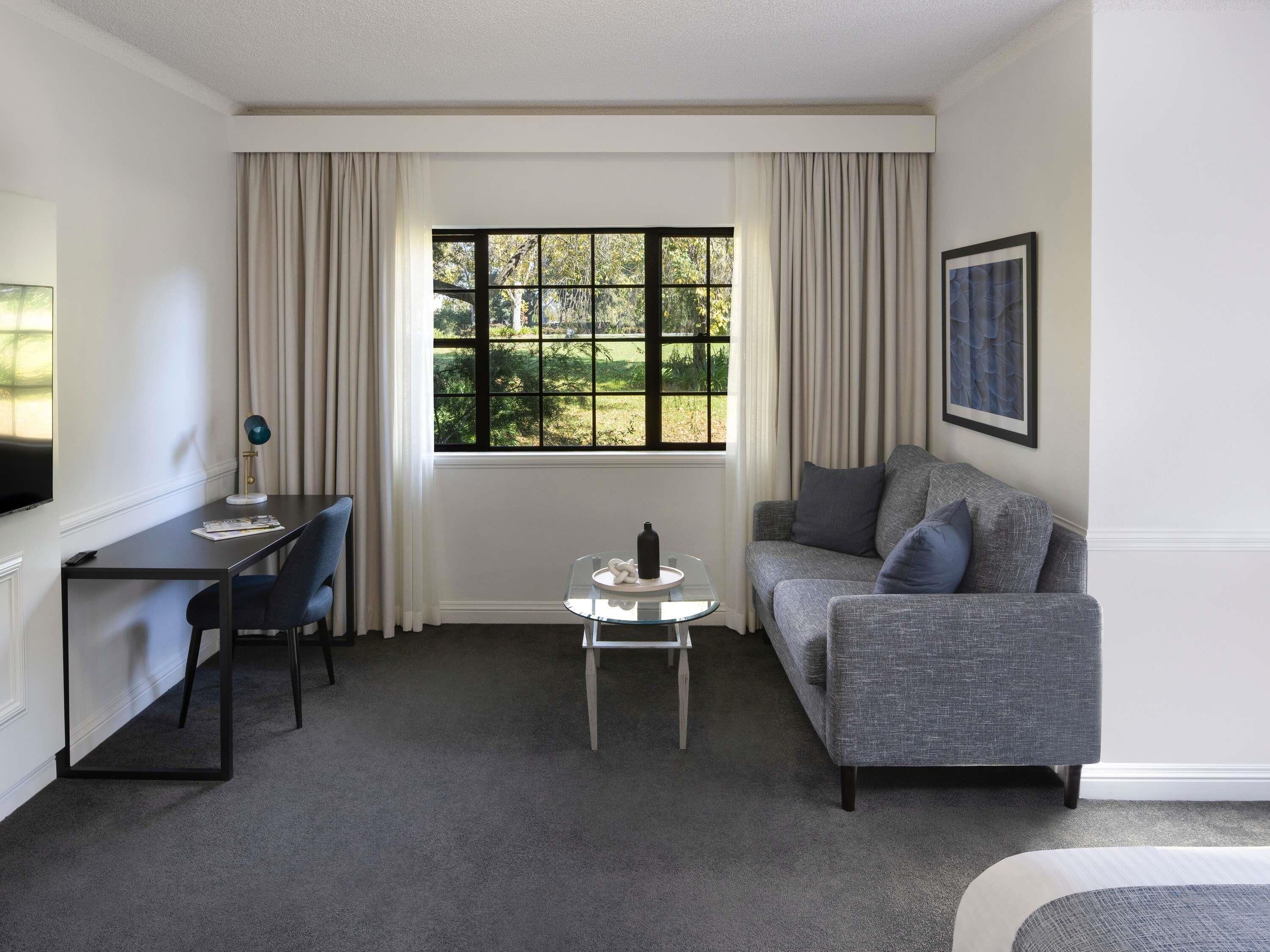A room with a grey sofa, a small table, a desk with a chair, and a window with curtains.
