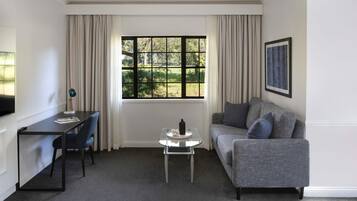 Una habitación con sofá gris, una mesita, un escritorio con silla y una ventana con cortinas.