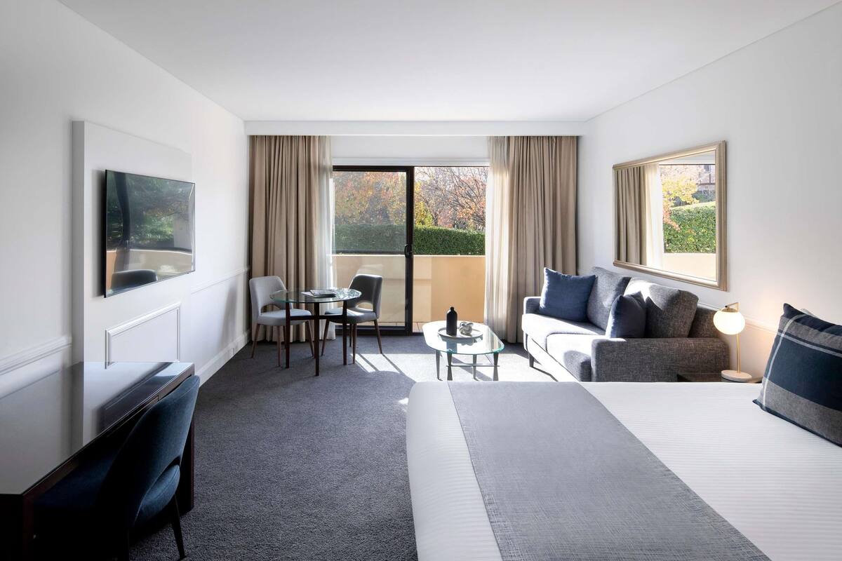 Ein Hotelzimmer mit Bett, Sofa, Schreibtisch, Stuhl, kleinem Tisch und an der Wand befestigtem Fernseher.