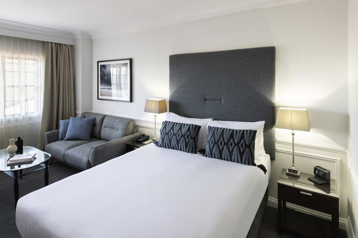 Ein Hotelzimmer mit einem großen Bett, einer Sitzecke, zwei Nachttischen, einer Lampe, einer Uhr und einem gerahmten Bild an der Wand.