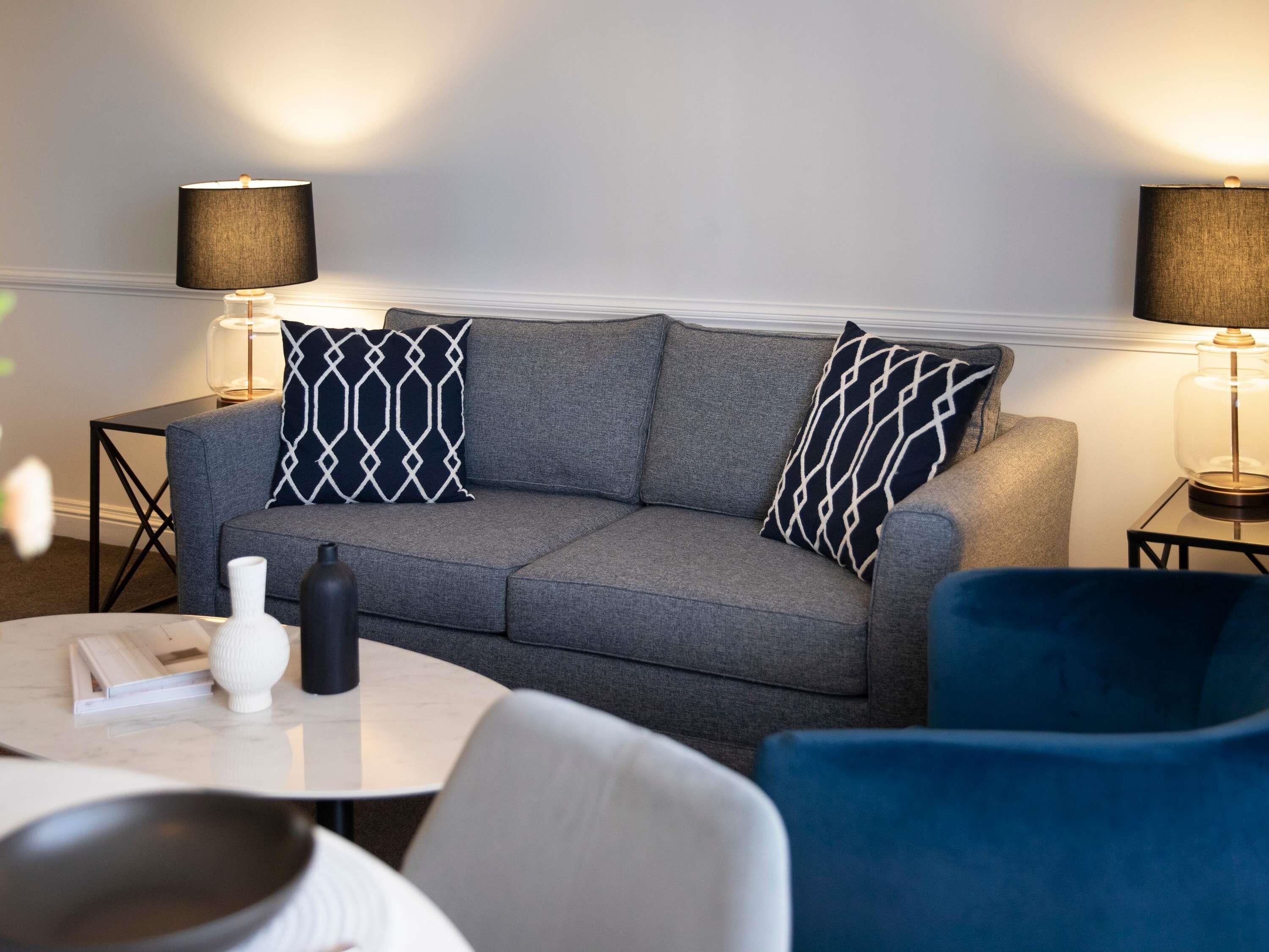 Un soggiorno moderno con un divano grigio, due cuscini decorativi a motivi, un vaso bianco, un vaso nero, una lampada in vetro e una poltrona blu.