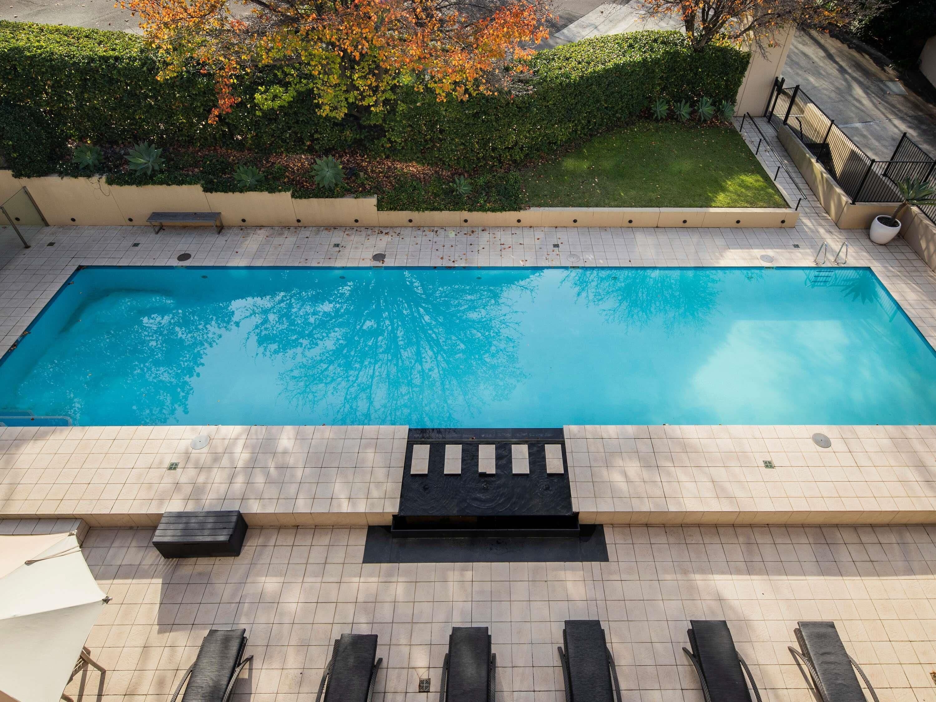 A rectangular swimming pool with a tiled deck, surrounded by a low hedge and trees with autumn foliage.