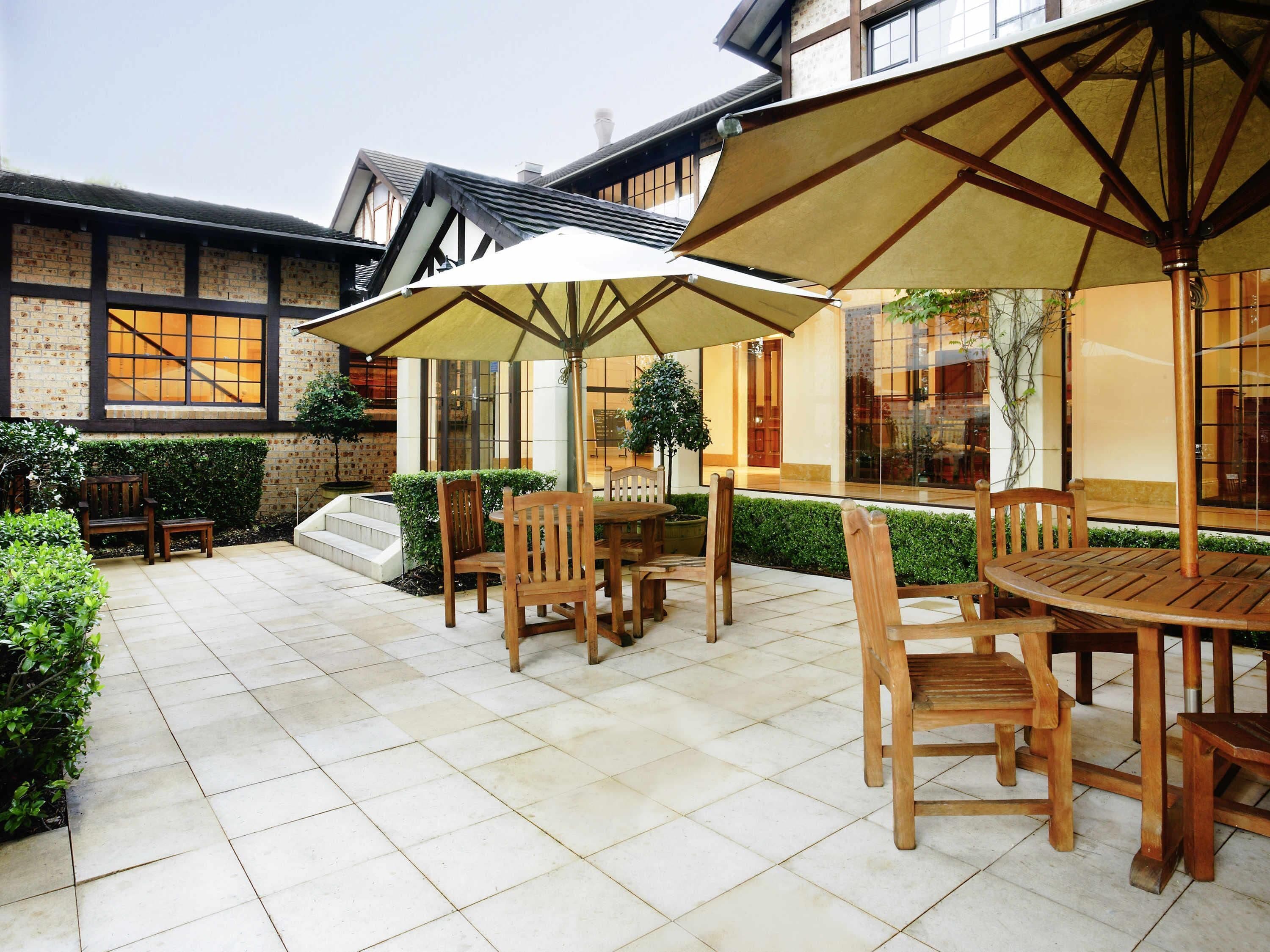 A patio area with wooden tables and chairs, large umbrellas, and a brick building in the background.