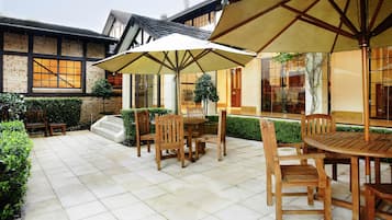 A patio area with wooden tables and chairs, large umbrellas, and a brick building in the background.