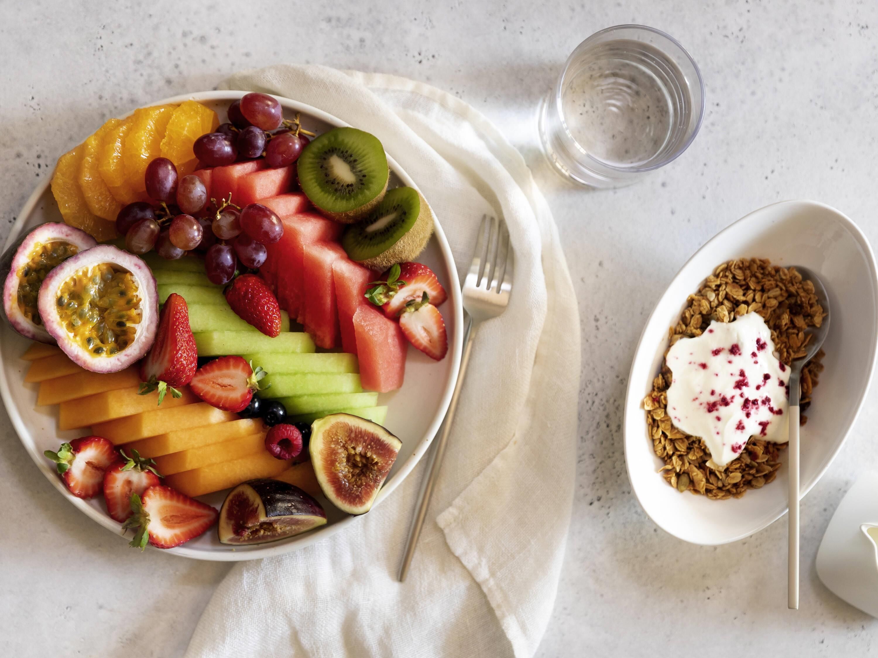 A fruit platter with various fruits and a bowl of granola with yogurt.
