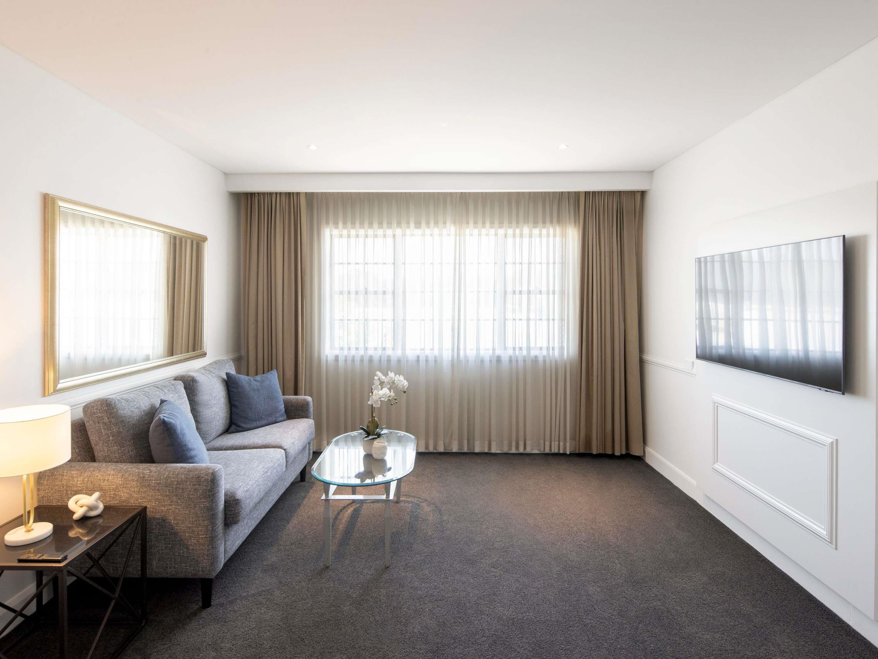 A modern hotel room with a grey sofa, a glass coffee table, a flat-screen TV, and a large window with curtains.