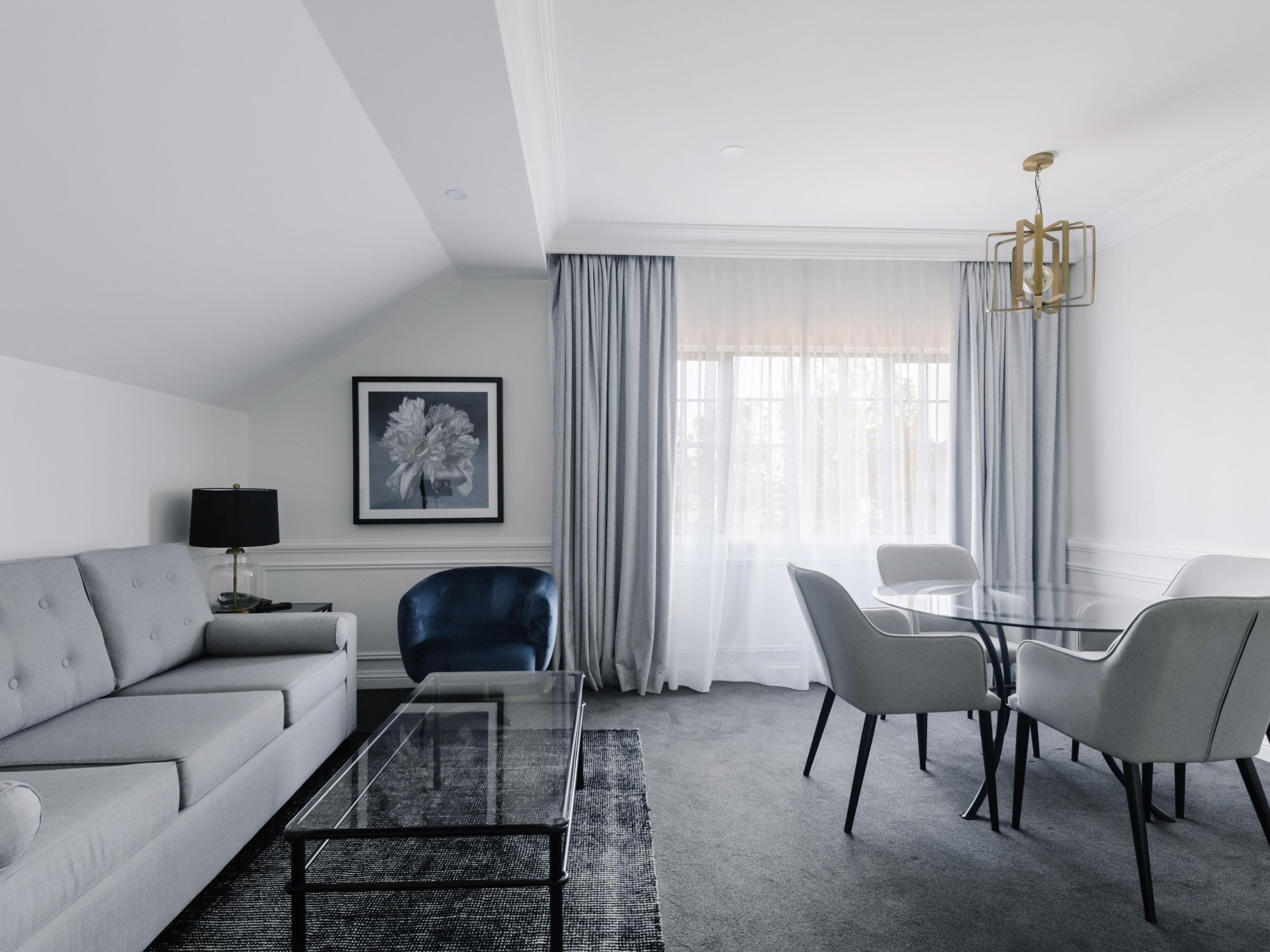 A modern living room with a sofa, a glass coffee table, a blue chair, a dining table with chairs, a chandelier, and a framed picture on the wall.