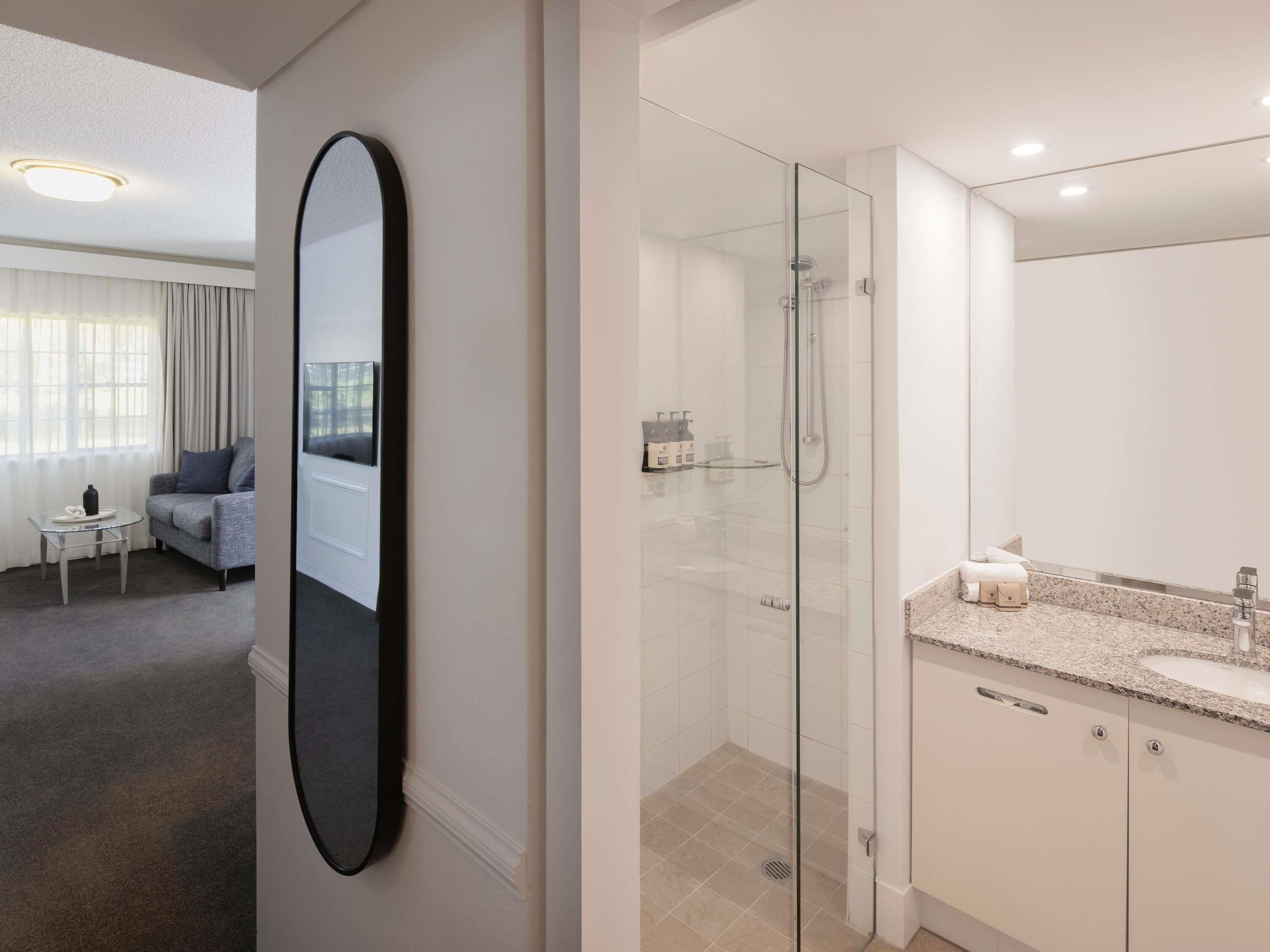 A modern hotel room with a glass-enclosed shower, a large mirror, a sofa, and a small table with a bottle and a glass.