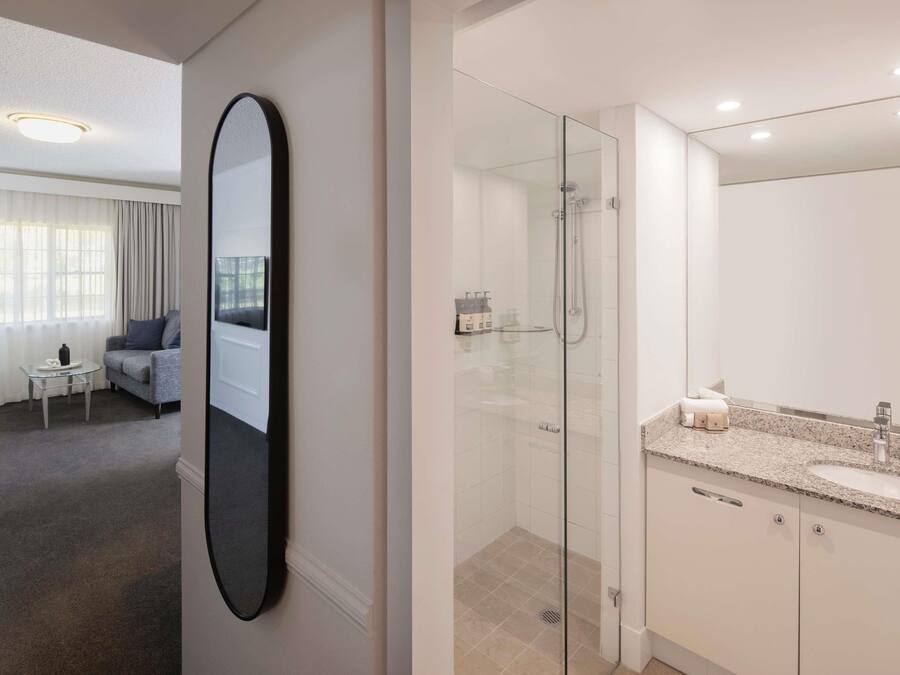 A modern hotel room with a glass-enclosed shower, a large mirror, a sofa, and a small table with a bottle and a glass.