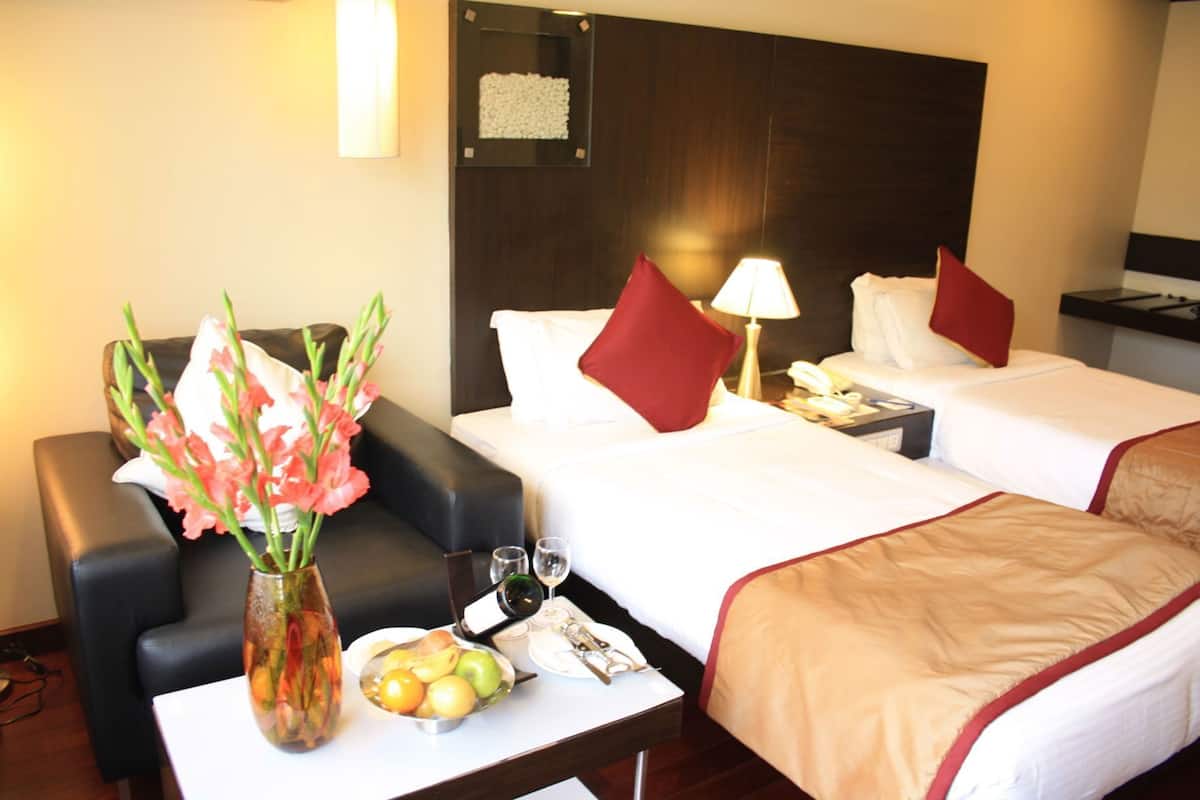 A hotel room with two beds, a sofa, a small table with a vase of flowers, and a bedside table with a lamp and a phone.