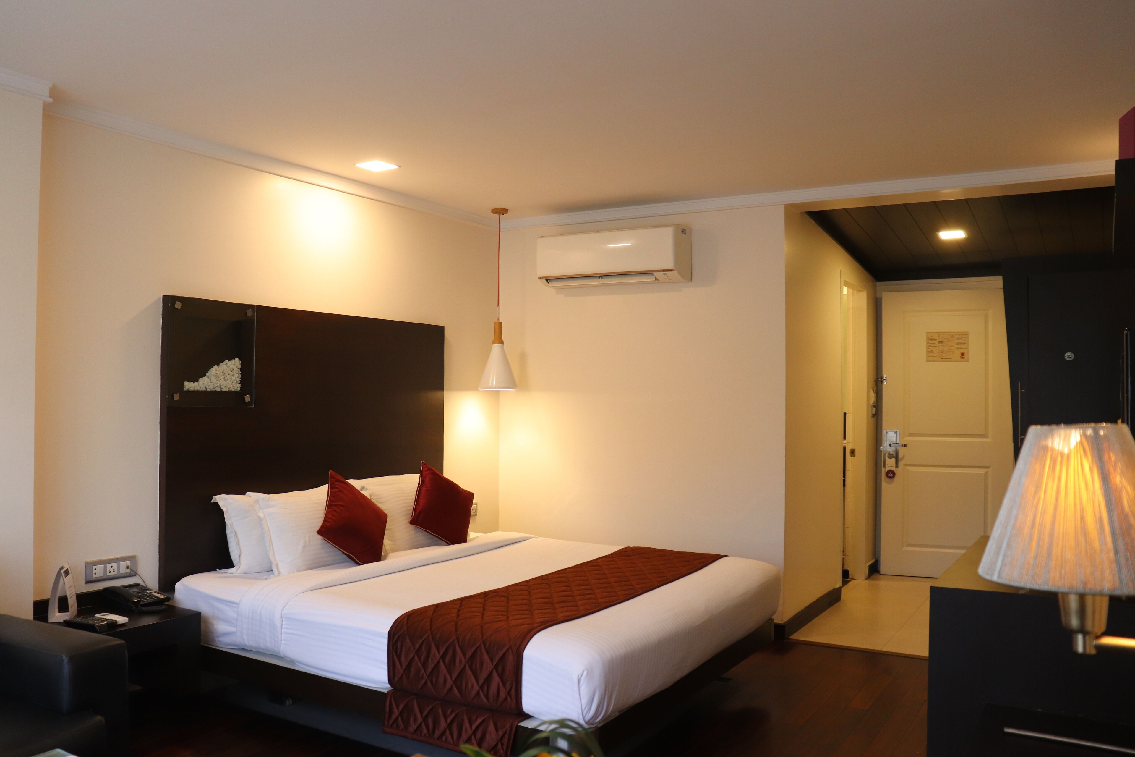 A hotel room with a bed, bedside table, lamp, and an air conditioning unit.