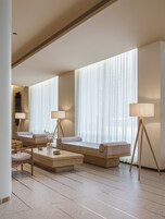 Una habitación de hotel moderna con un sofá, dos lámparas de pie, una mesita de centro y un ventanal con cortinas traslúcidas.
