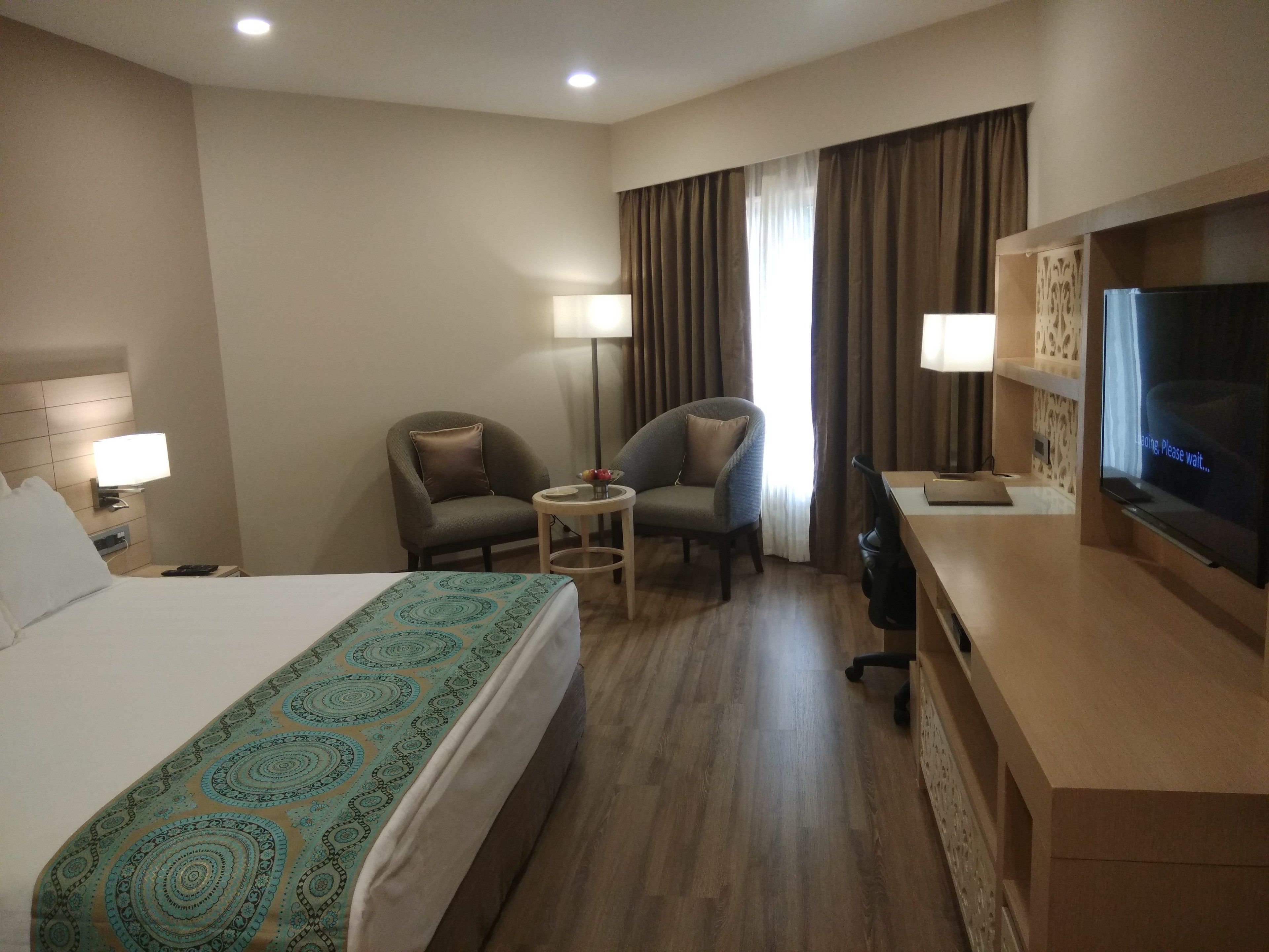 A hotel room with a bed, two armchairs, a small table, a desk with a computer, a TV, and a window with curtains.
