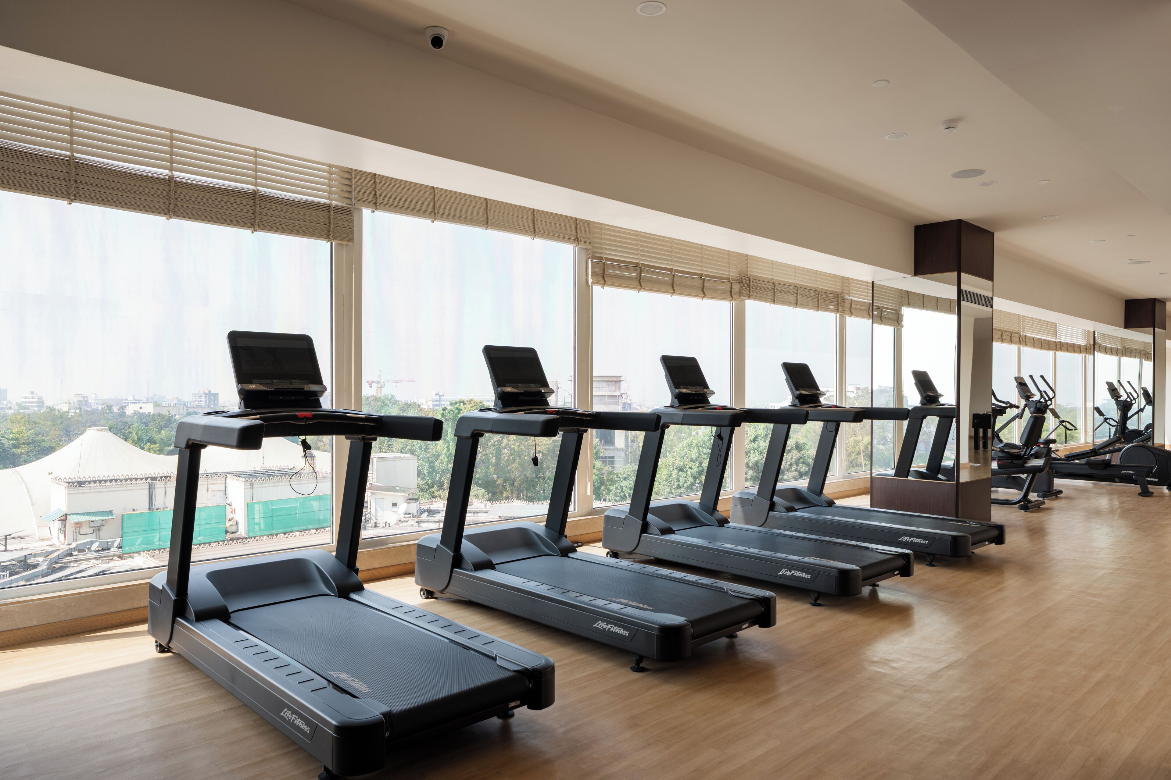 Un gimnasio con cintas de correr y bicicletas estáticas, ventanas amplias con persianas y vistas a edificios exteriores.