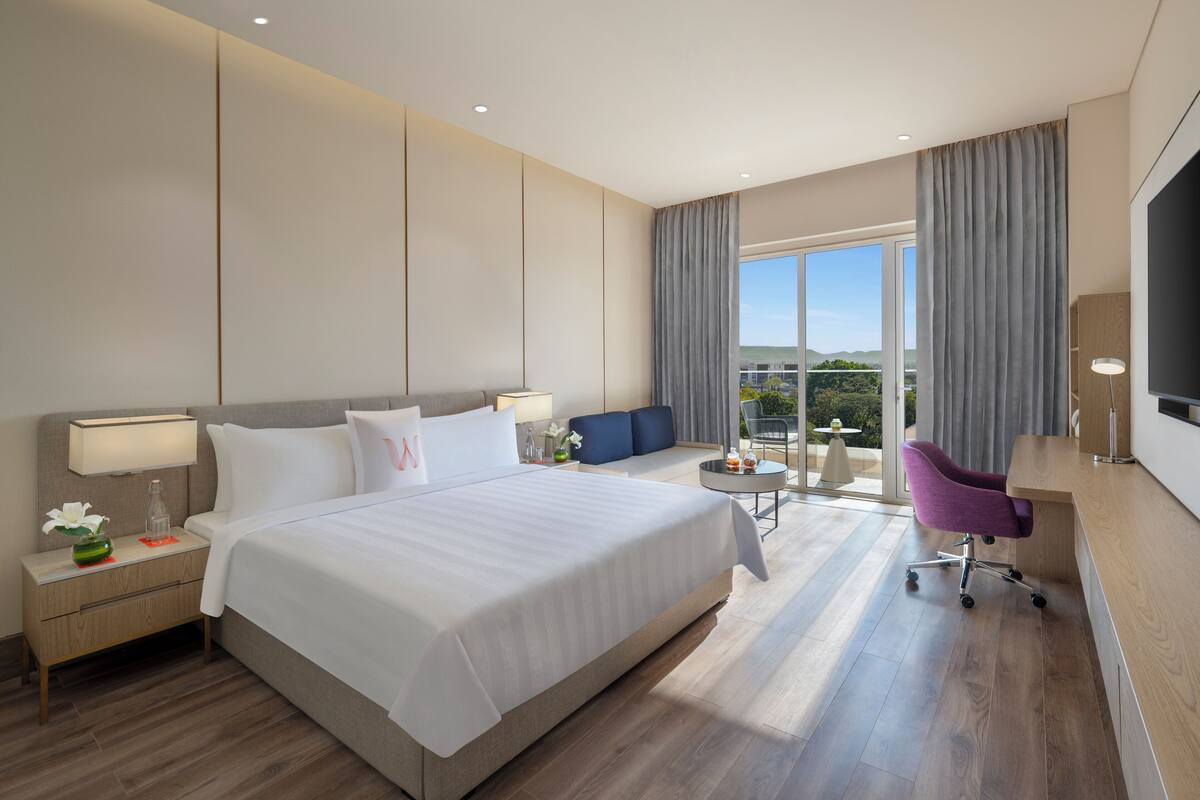 Ein modernes Hotelzimmer mit einem großen Bett, einem Schreibtisch, einem Sessel und Blick ins Freie.