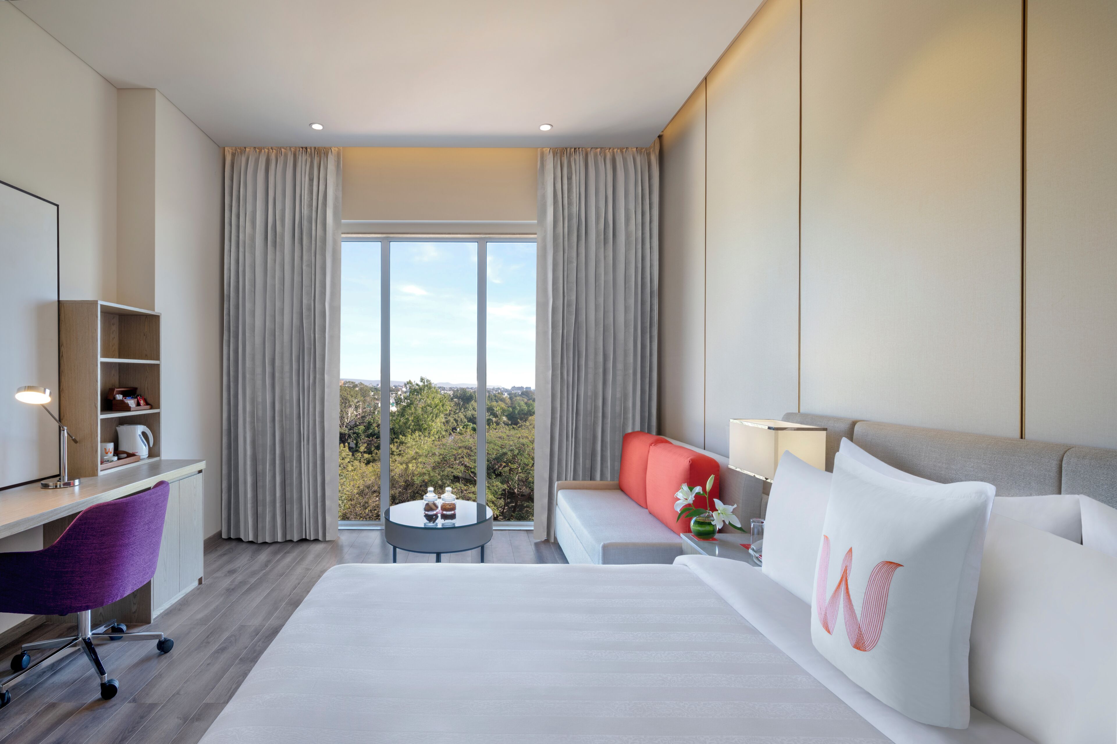 A hotel room with a bed, a desk, a chair, a sofa, a small table, and a view of trees through large windows.