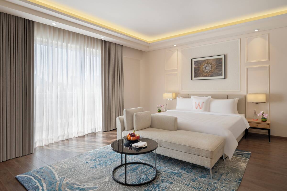 Ein modernes Hotelzimmer mit einem großen Bett, einer Couch, einem kleinen Tisch mit Früchten und einem großen Fenster mit Vorhängen.