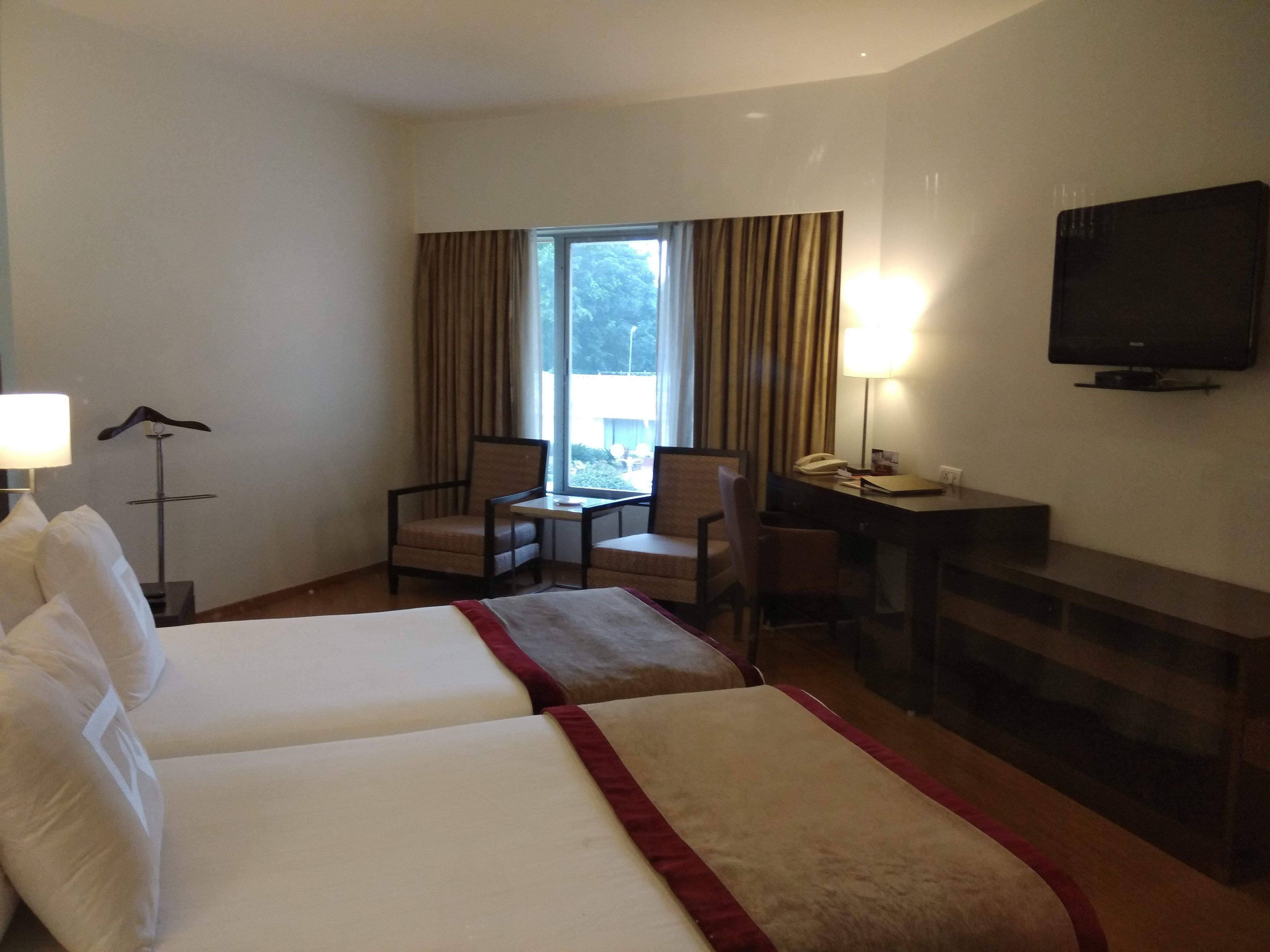 A hotel room with two beds, a desk, a chair, a lamp, a TV, and a window with curtains.