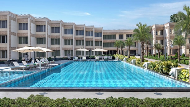 Une piscine entourée de chaises longues et de parasols, elle-même bordée par un bâtiment d’hôtel à plusieurs étages doté de balcons.