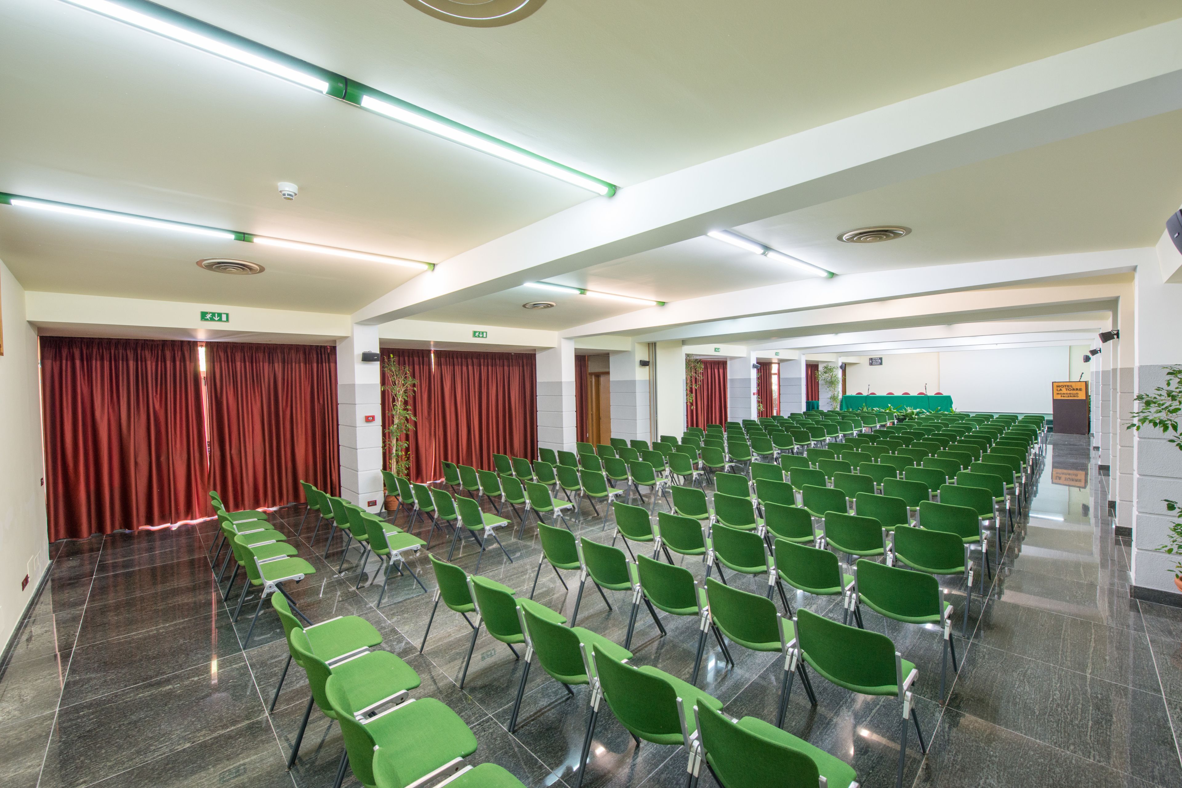 A conference room with rows of green chairs, a stage with a red curtain, and a green exit sign.