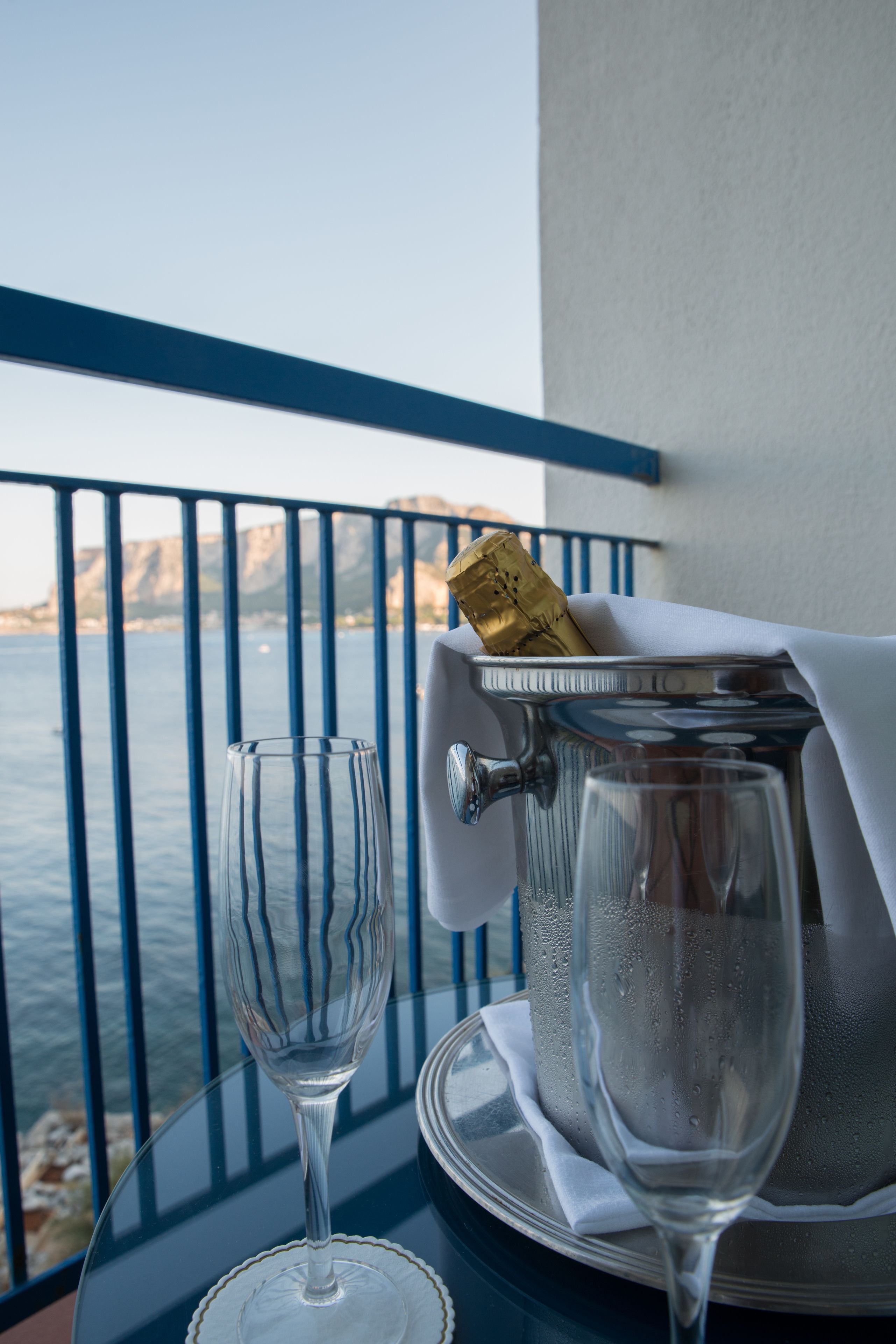 A glass of water, a bottle of champagne, and a glass decanter on a table with a view of the sea and cliffs.