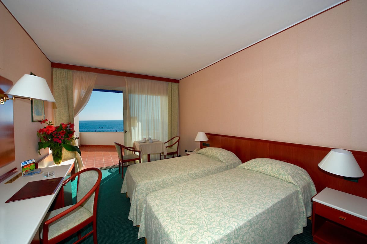 A hotel room with two beds, a desk, a chair, a table, and a view of the sea.