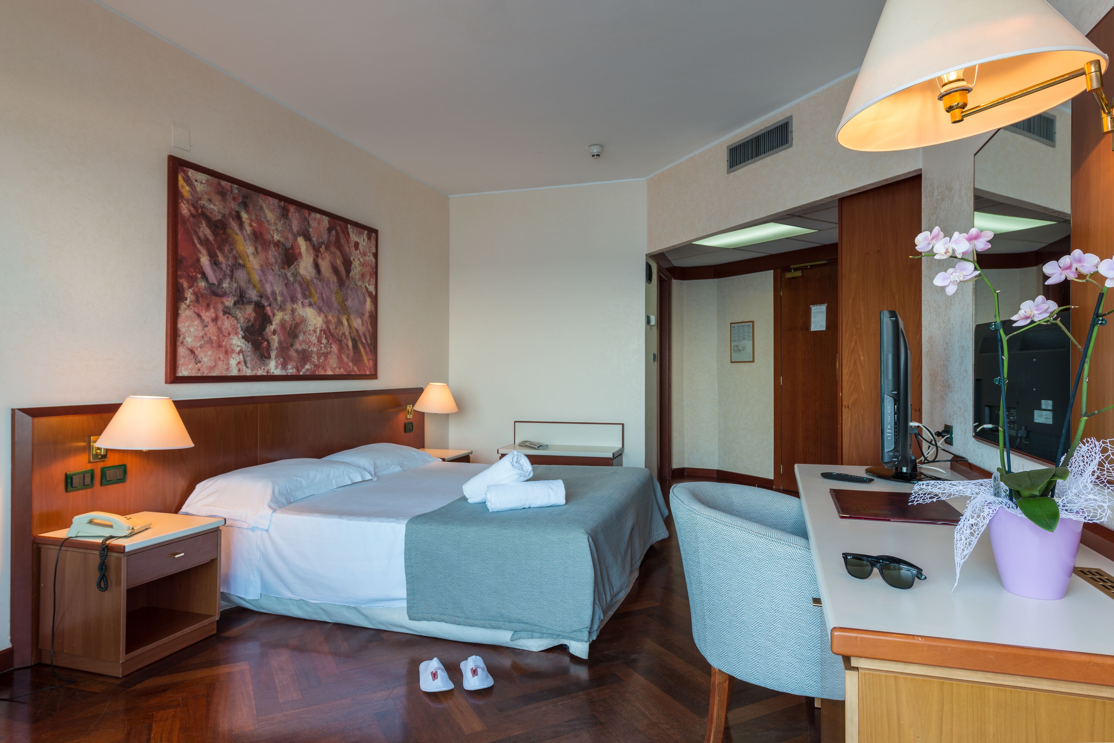 Une chambre d’hôtel avec un lit, des lampes de chevet, un bureau avec un miroir, une chaise et une plante en pot.