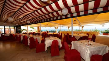 A spacious outdoor dining area with multiple tables and red chairs, under a striped canopy, overlooking a scenic view.