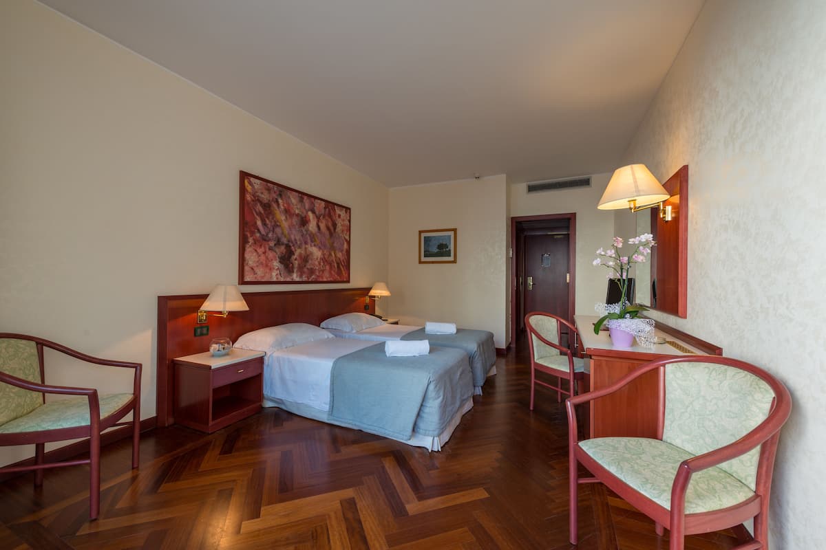 A hotel room with two beds, wooden flooring, a desk with a lamp, and two chairs.