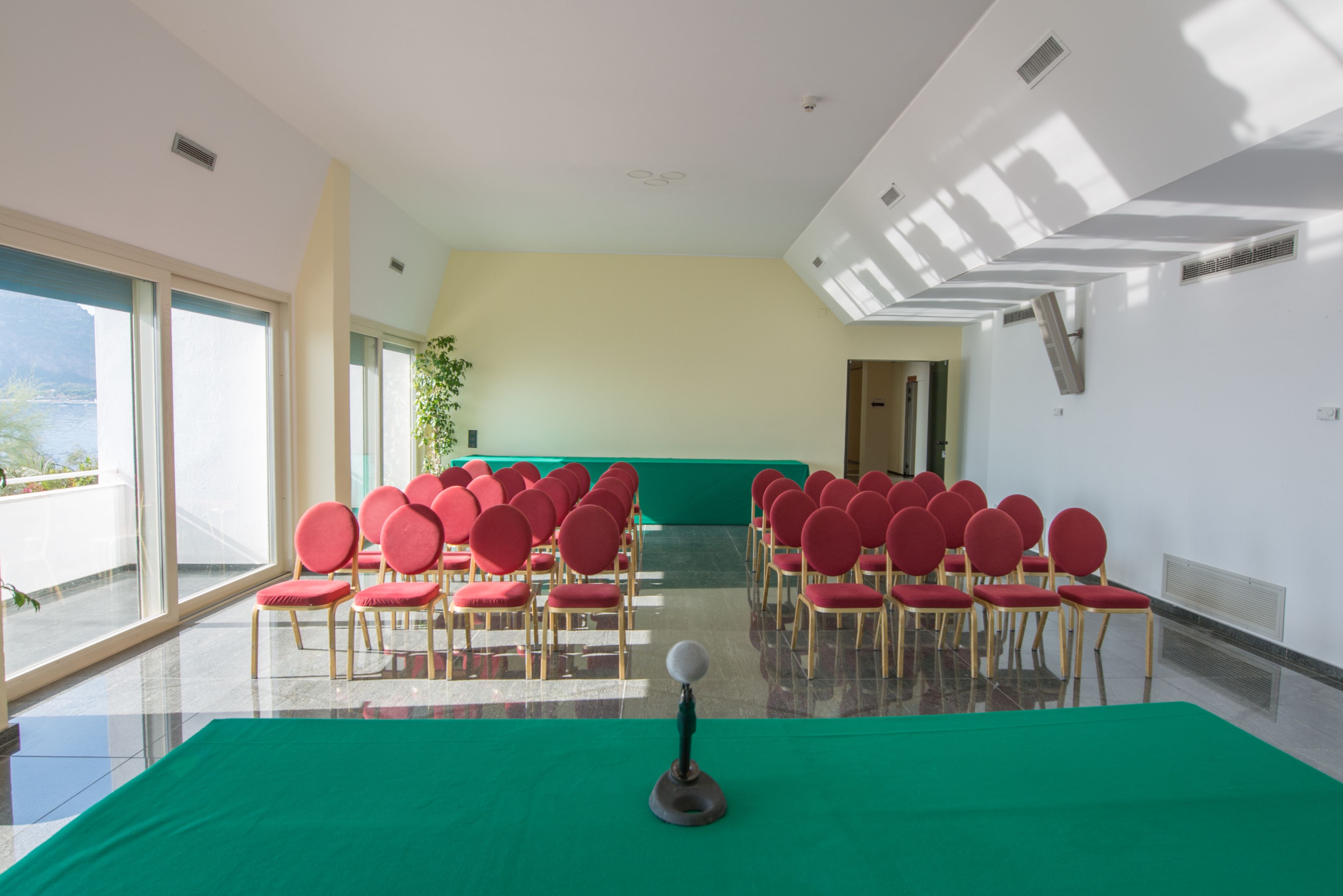 Een vergaderzaal met rijen rode stoelen, een groene tafel en een groot raam met uitzicht op het omliggende landschap.