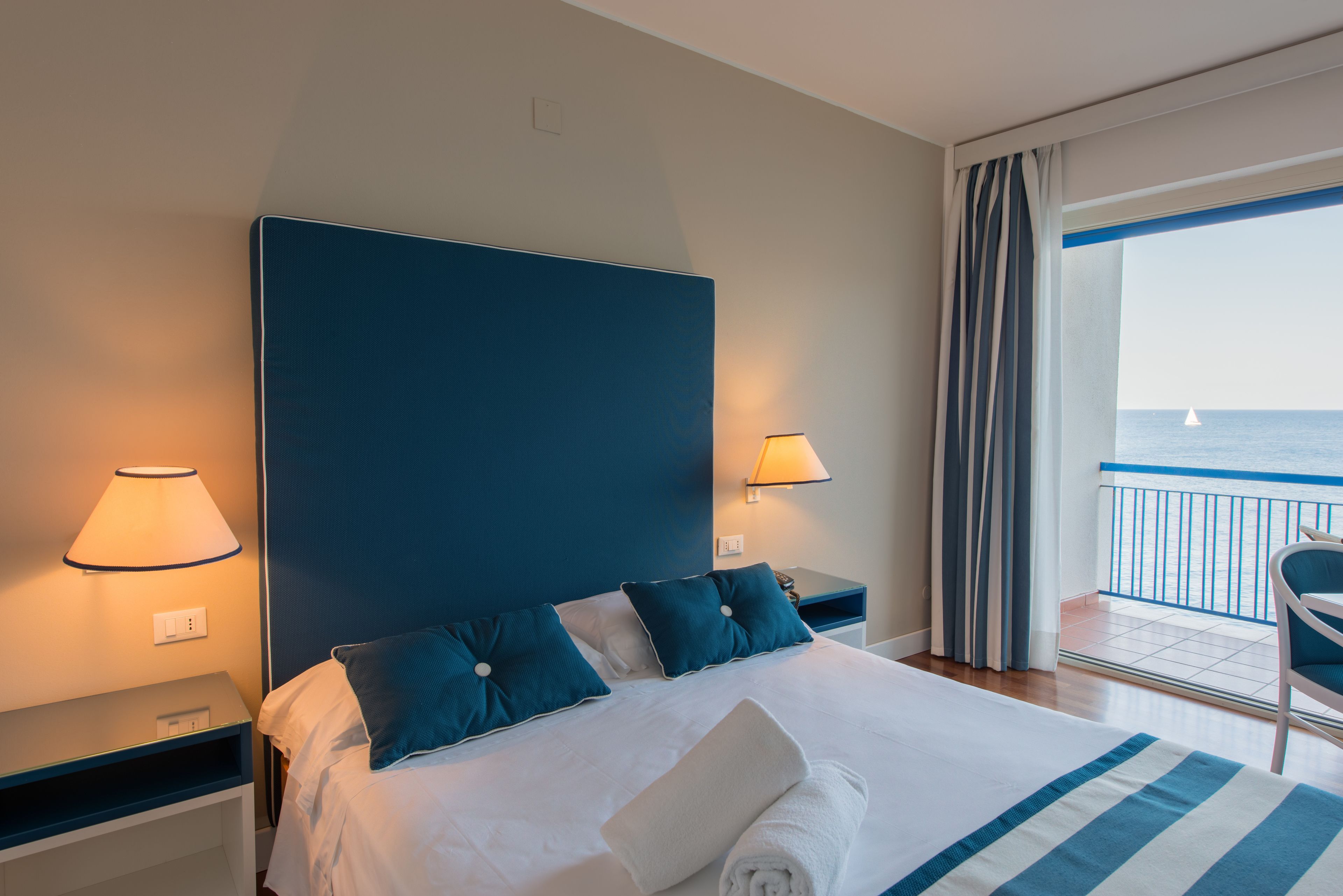 Une chambre d’hôtel avec un grand lit, deux tables de chevet avec des lampes et une vue sur la mer depuis un balcon.