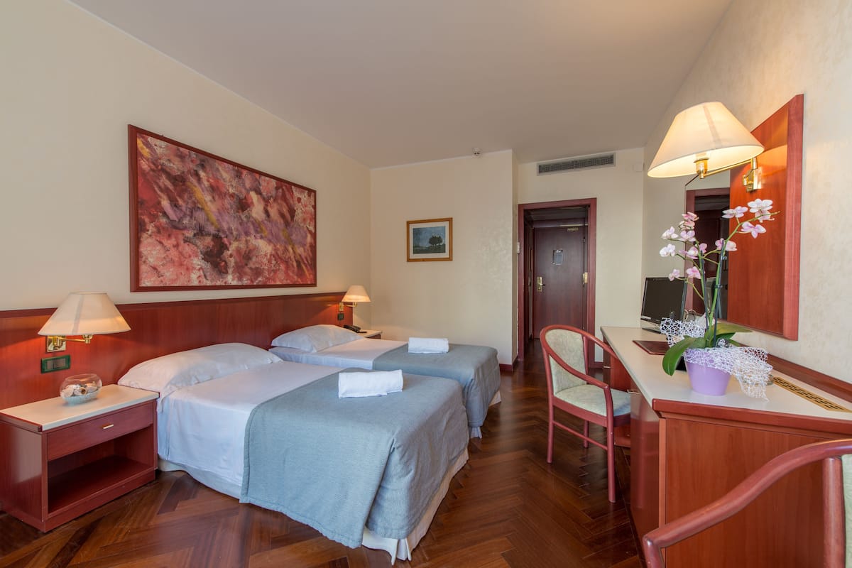 A hotel room with two beds, a desk, a chair, a lamp, and a painting on the wall.