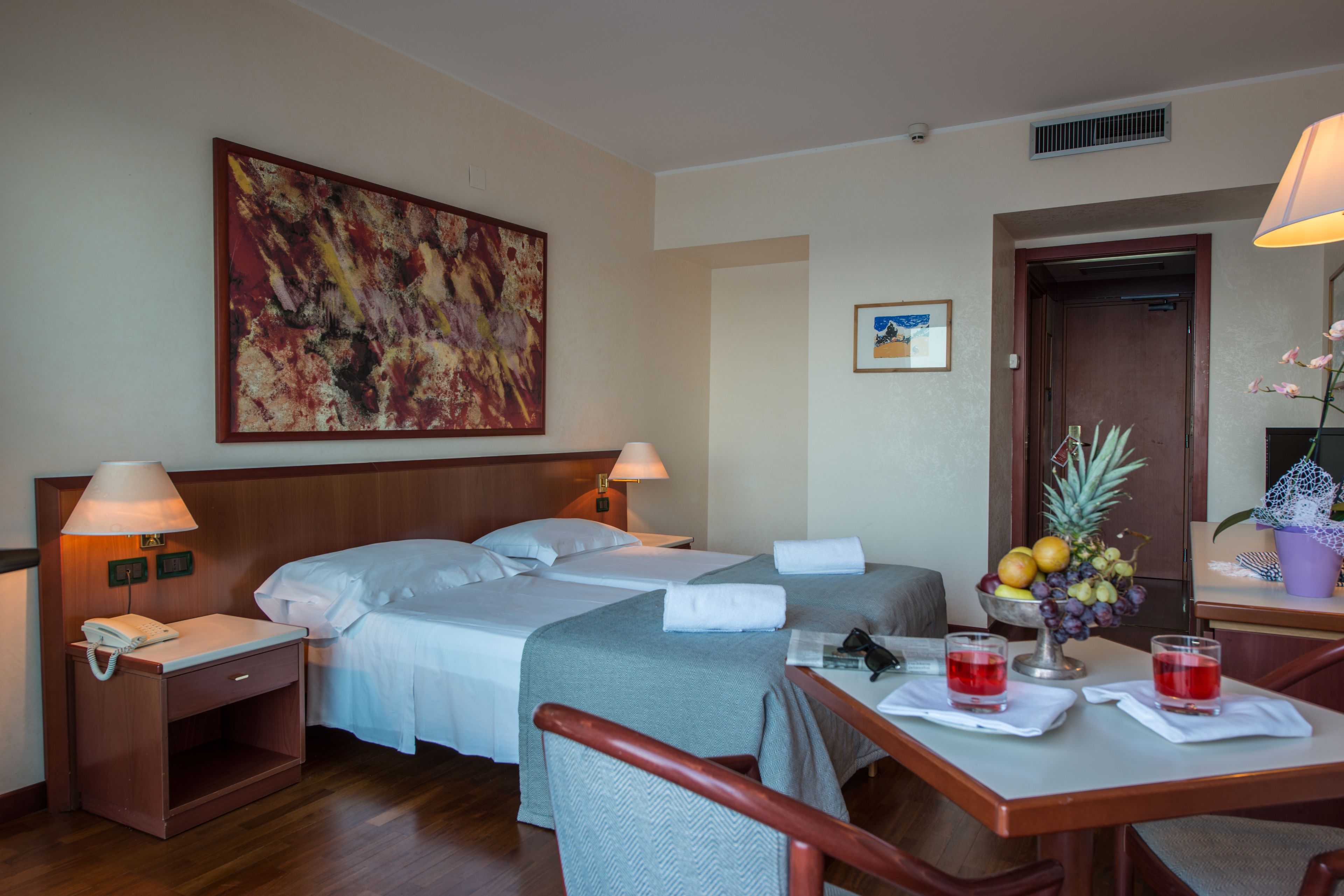 Une chambre d’hôtel avec deux lits, un bureau sur lequel se trouve un panier de fruits, et un tableau accroché au mur.