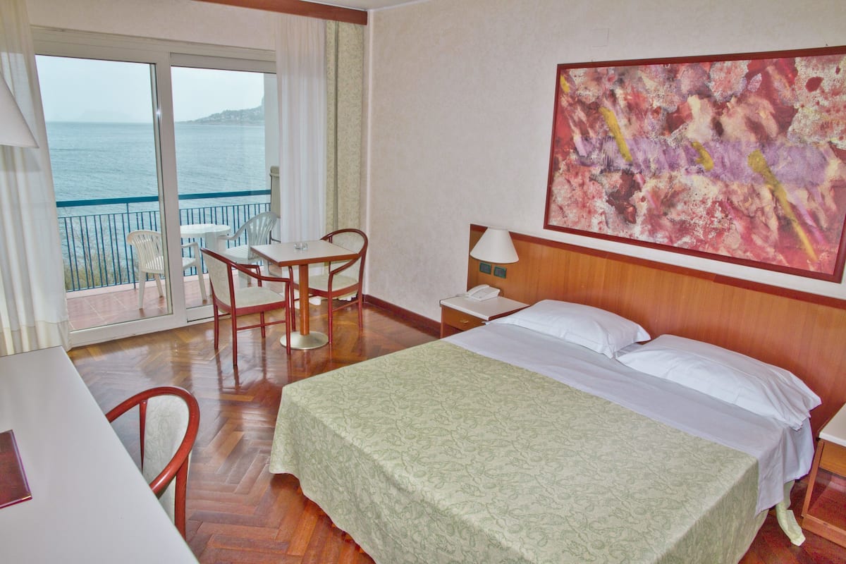 A hotel room with a bed, a desk with a chair, a table, a lamp, and a painting on the wall.