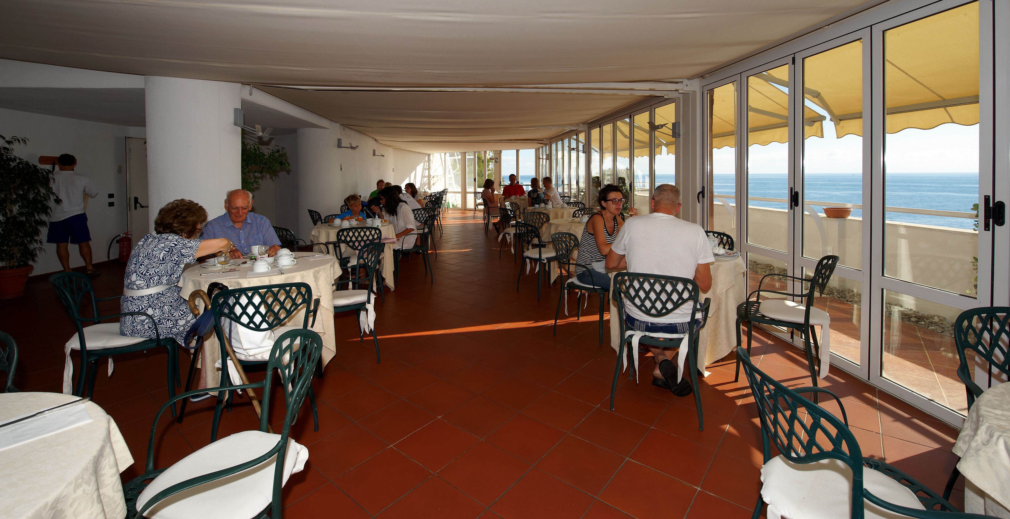 Un'area pranzo all'aperto coperta, con tavoli e sedie, ampie finestre e vista sul mare.