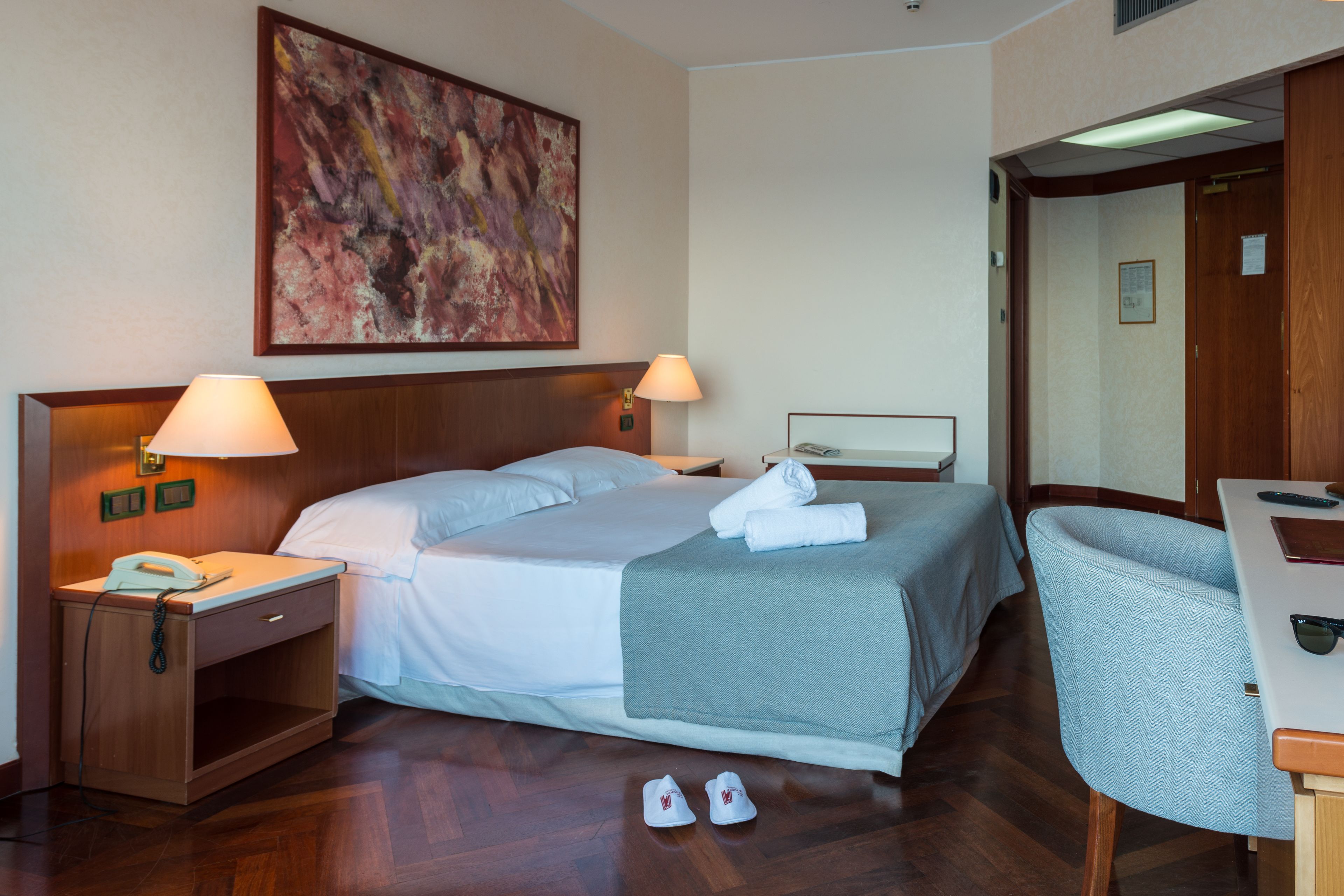 Una camera d'albergo con un letto, due lampade ai lati del letto, una scrivania con il telefono, una sedia e un paio di scarpe sul pavimento.