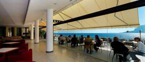 Un área de comedor al aire libre con mesas y sillas, una sombrilla grande que proporciona sombra, y vista al mar.