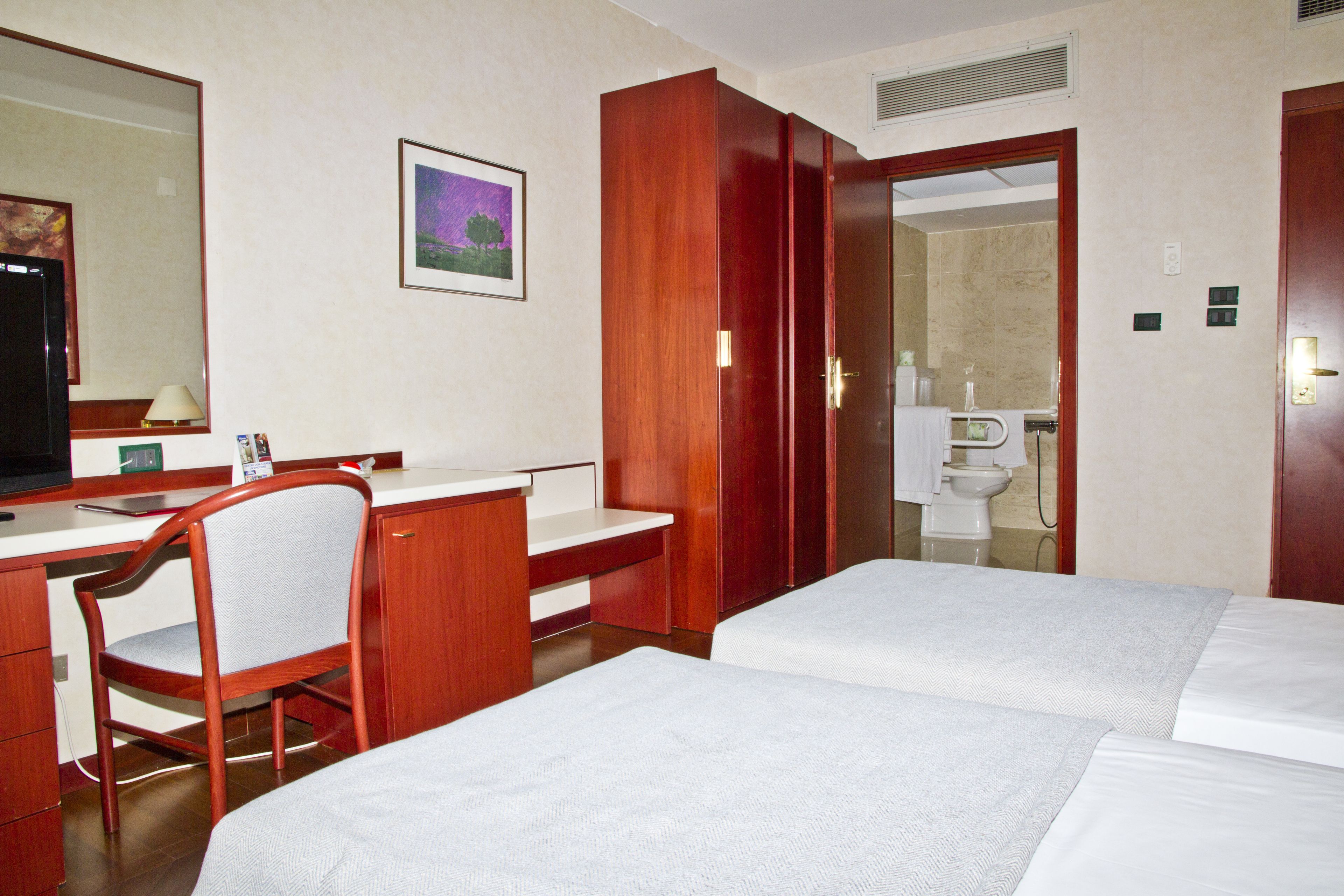 A hotel room with a desk, two beds, a chair, a mirror, and a bathroom visible through an open door.