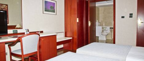 A hotel room with a desk, two beds, a chair, a mirror, and a bathroom visible through an open door.