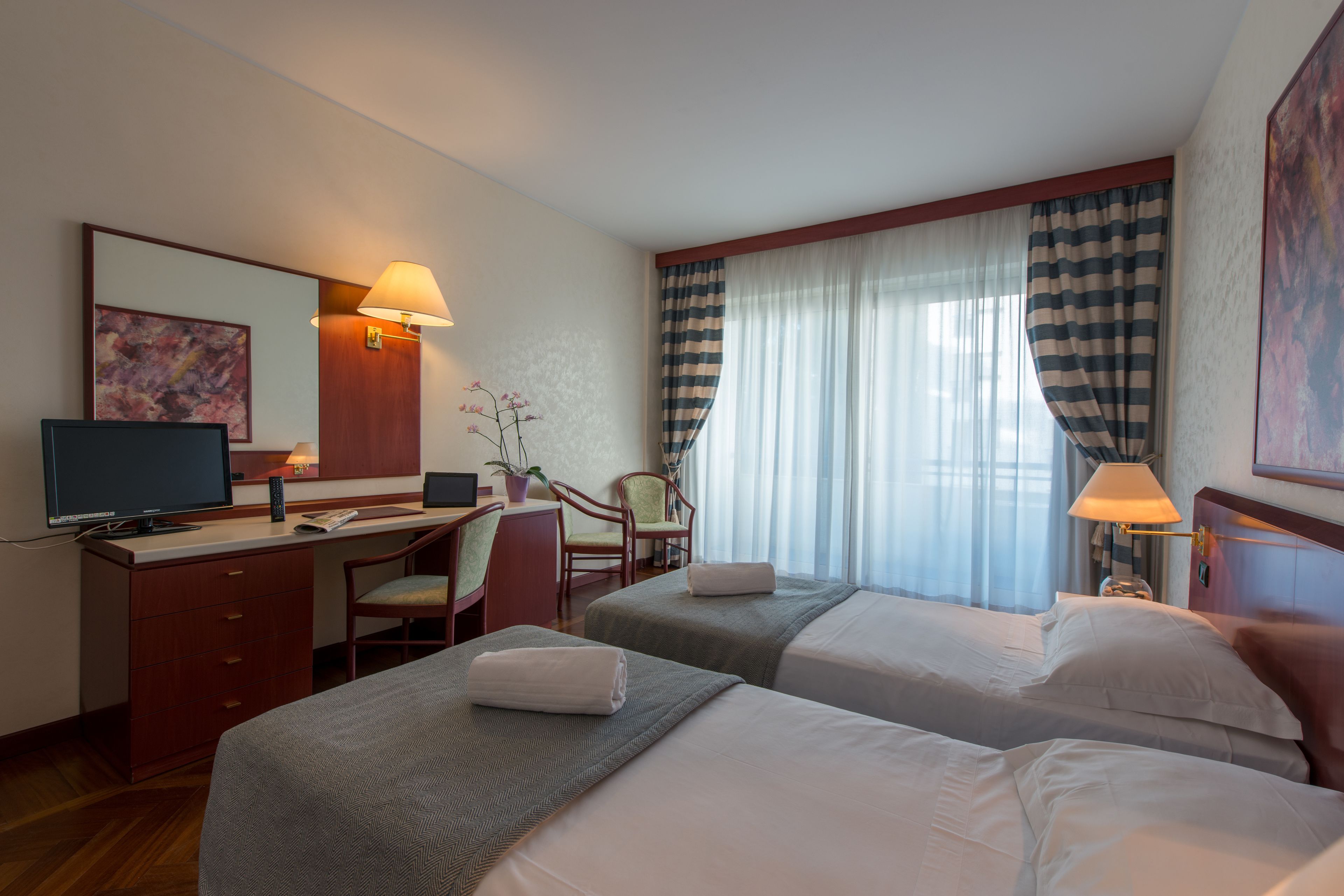 Une chambre d’hôtel avec deux lits, un bureau, une chaise, une télévision et une grande fenêtre avec des rideaux.