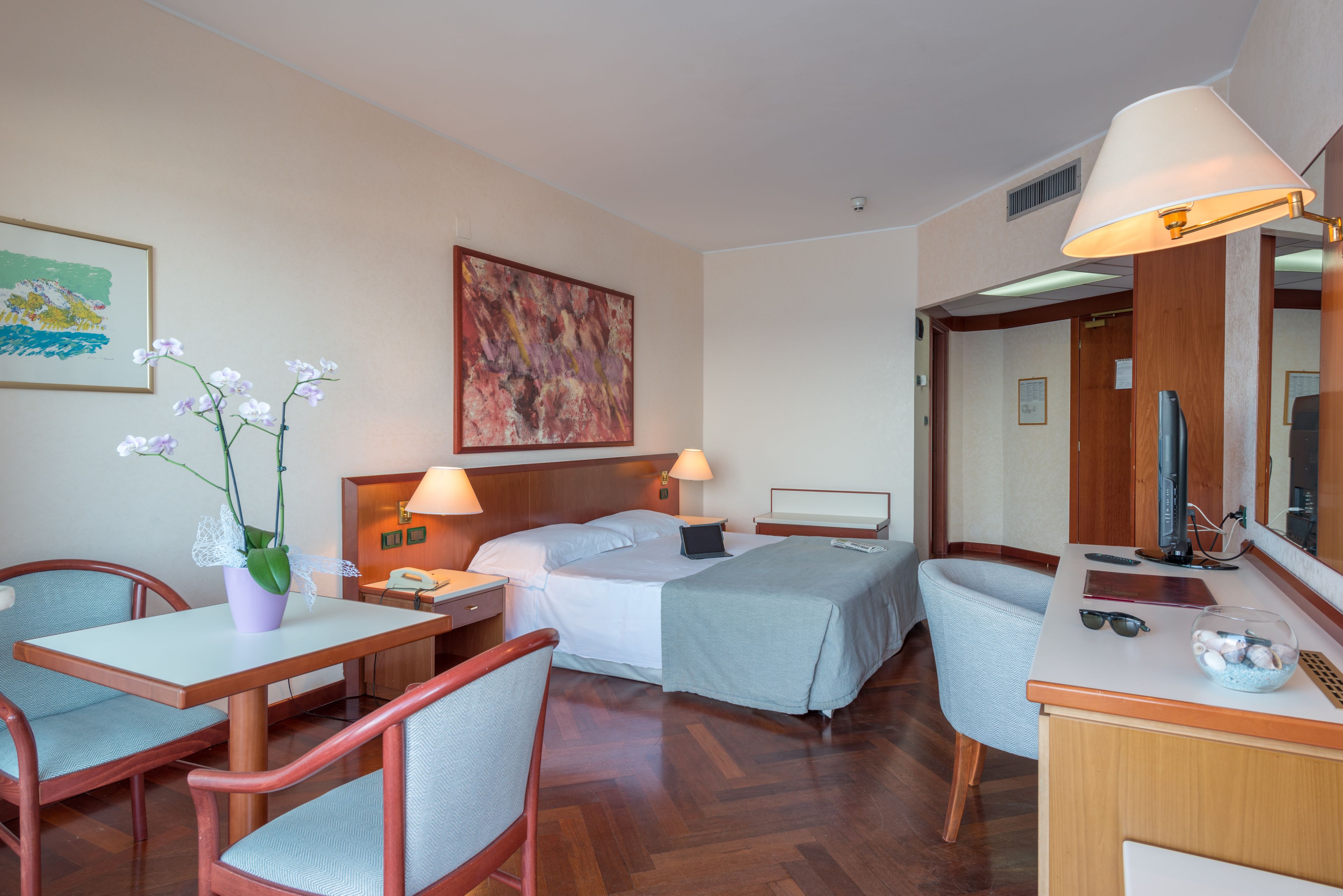 Habitació d'hotel amb un llit, taules de nit, escriptori i cadira, taula petita amb un vas de flors, làmpada i mirall.