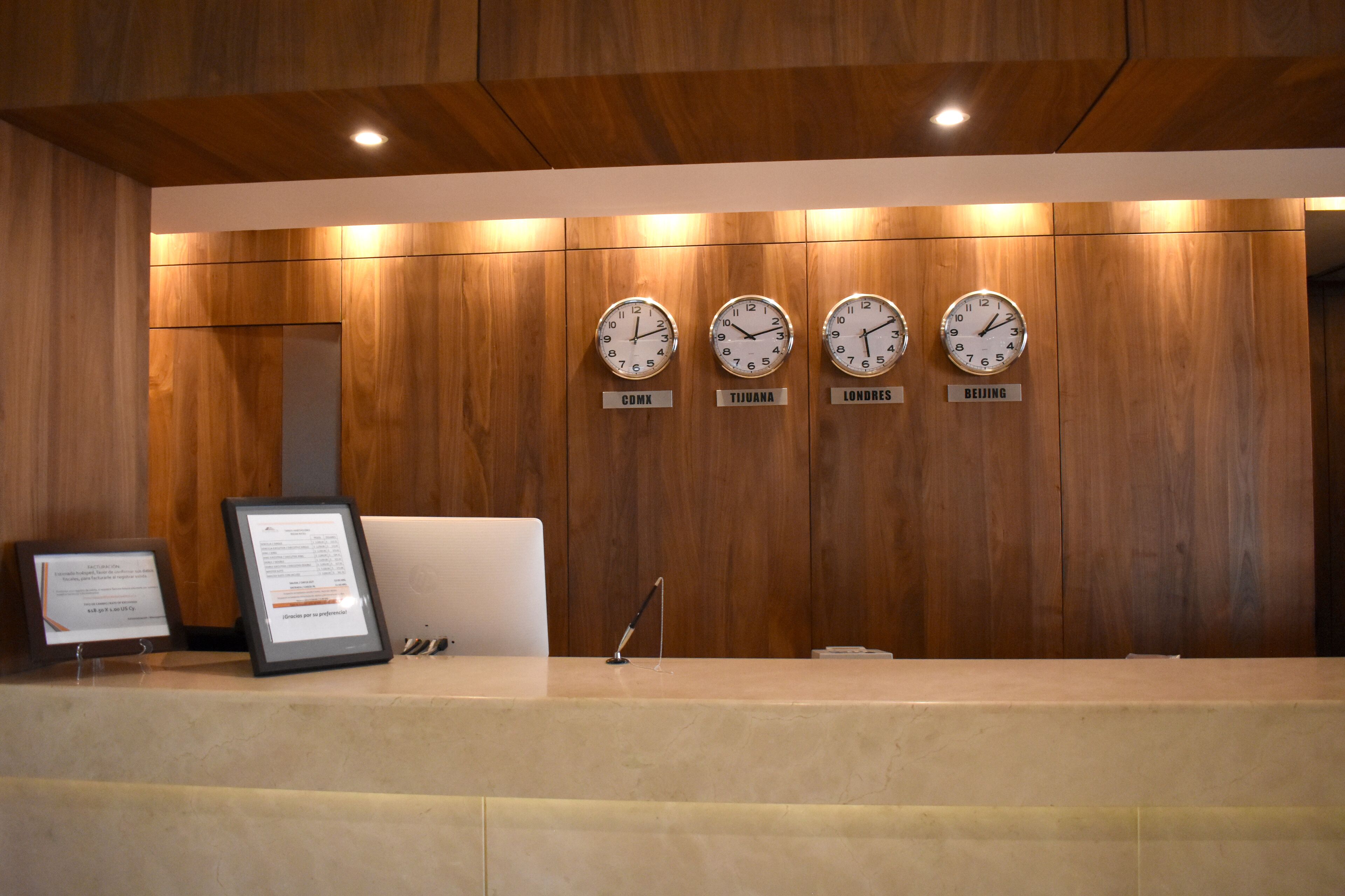 Un mostrador de recepción con relojes mostrando horarios de distintas ciudades, carteles informativos enmarcados y un interior de madera moderna.