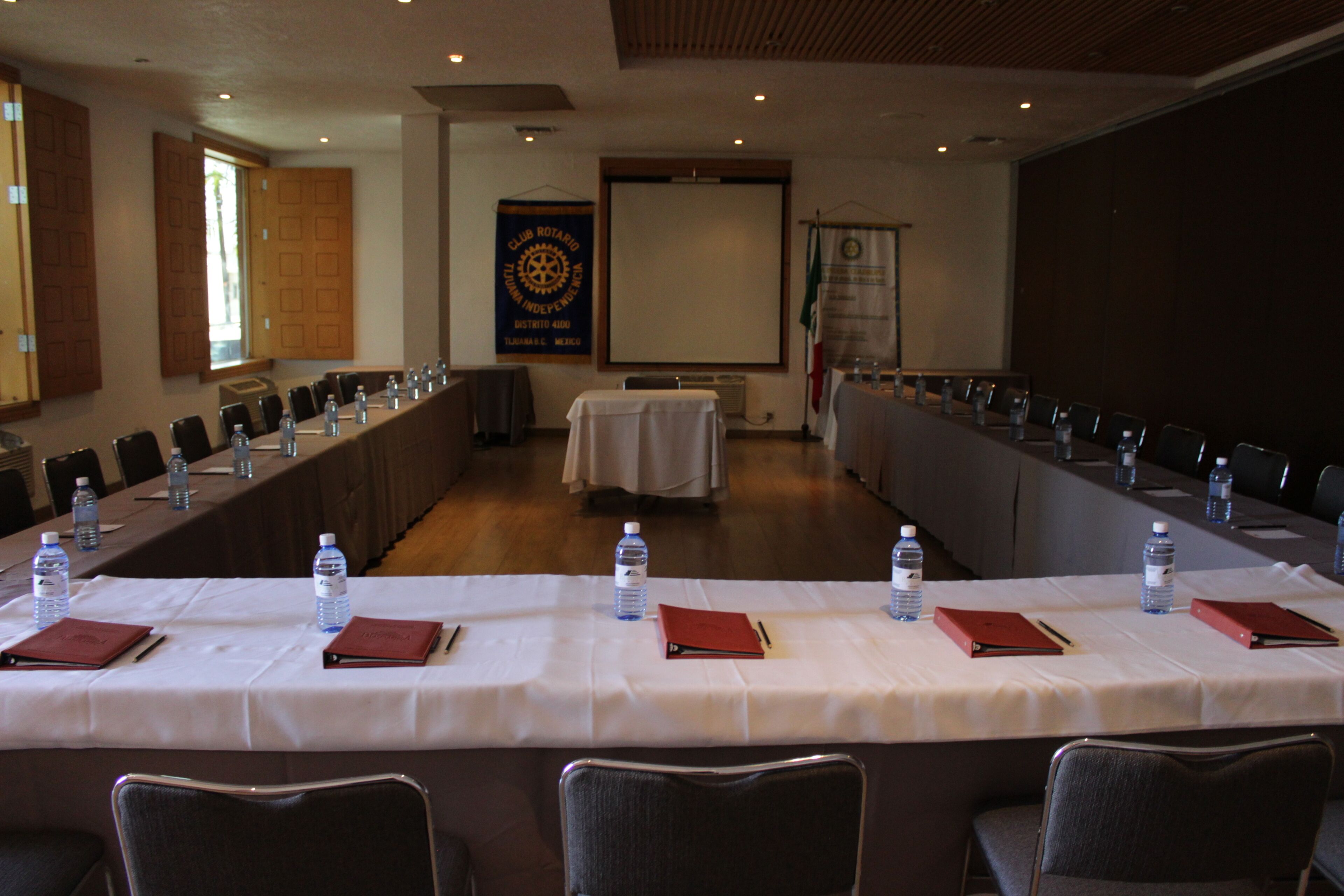 Un salón de conferencias con una larga mesa, sillas, botellas de agua y una pantalla de proyector.