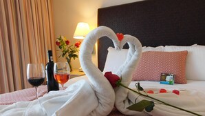 A neatly made bed with a heart-shaped towel arrangement, a bottle of wine, and a rose on the bed.